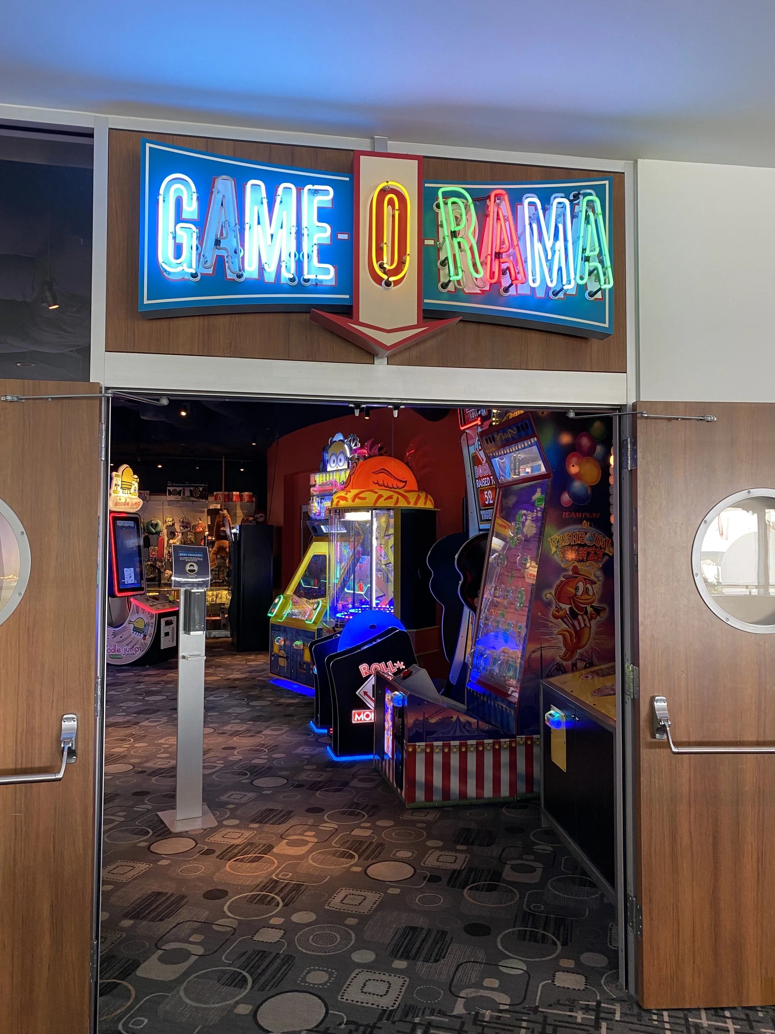 Entrance to an arcade with a neon sign that reads 'GAME-OF-RAMA'. Inside the arcade, there are various colorful arcade machines and games.