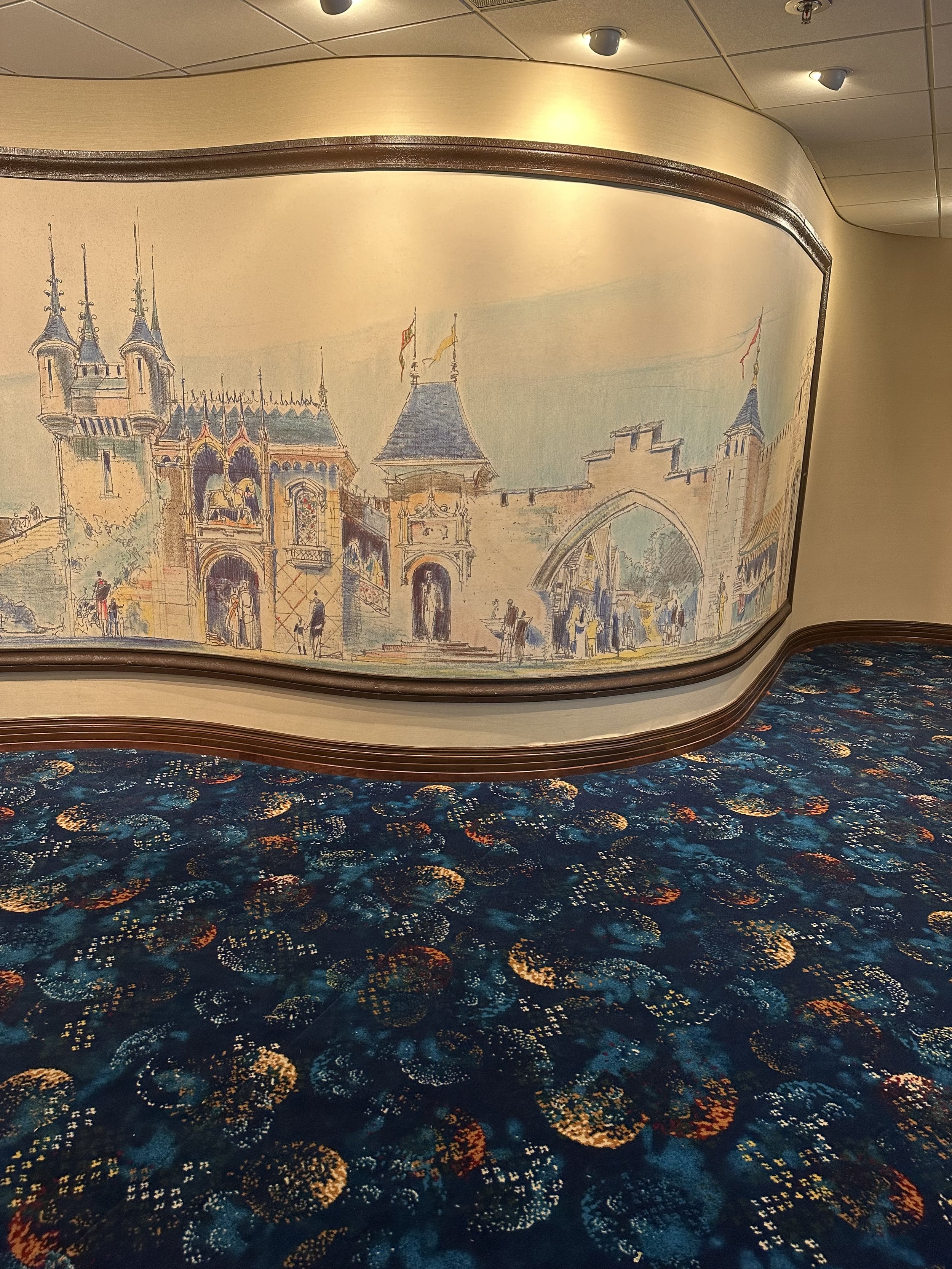 Illustration of a castle with towers and archways, painted on a wall, behind a curved wooden molding, with a carpeted floor featuring a colorful, circular pattern.