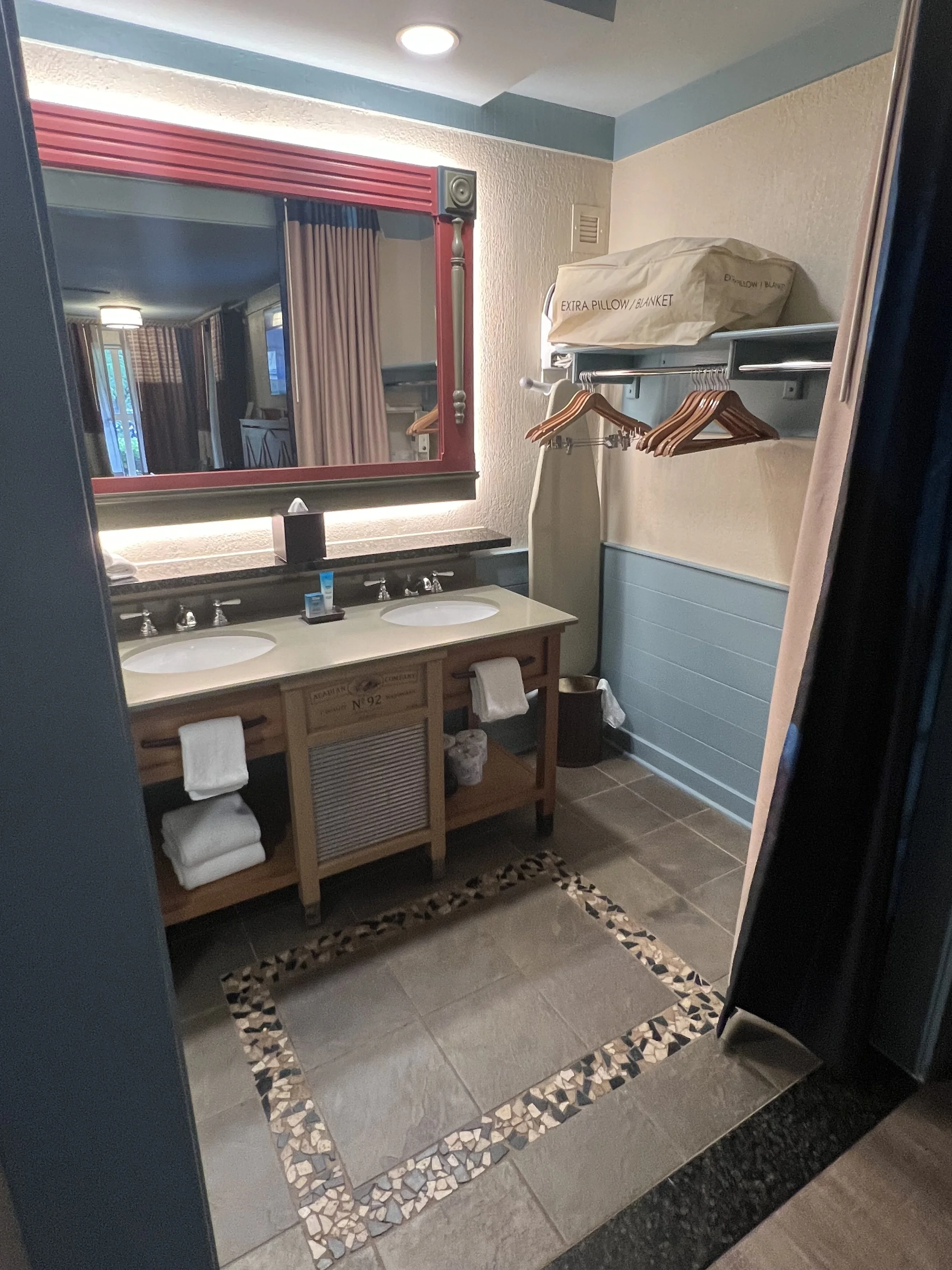 Bathroom with double sinks, large mirror, and a window with curtains. There are towels, an ironing board, and hanging space for clothes. The floor has a decorative tile border.