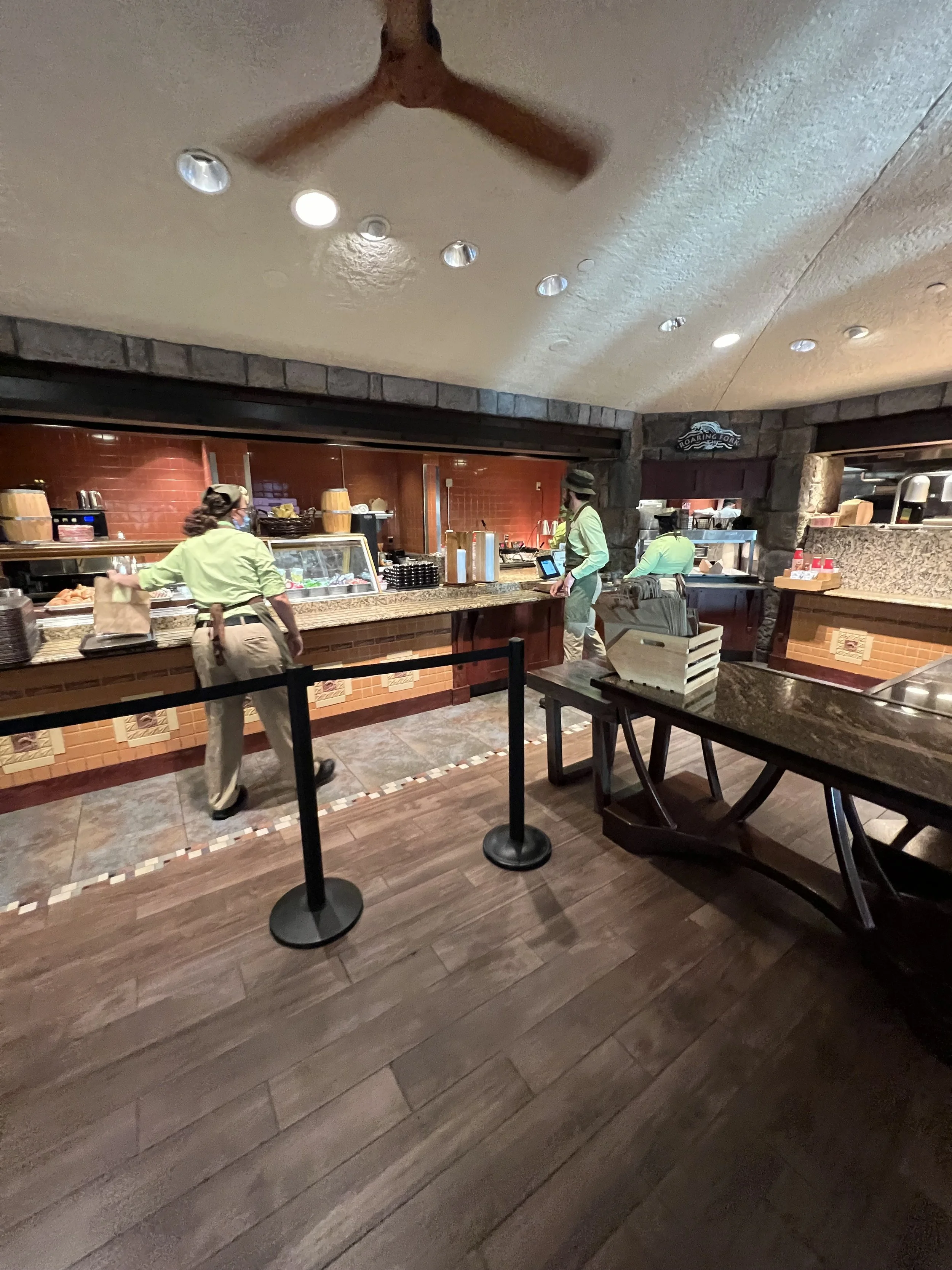 Inside a restaurant, three staff members wearing green uniforms and hats work behind a counter with warm lighting, a glass display case, and utensils, while a table is pushed aside in the foreground.