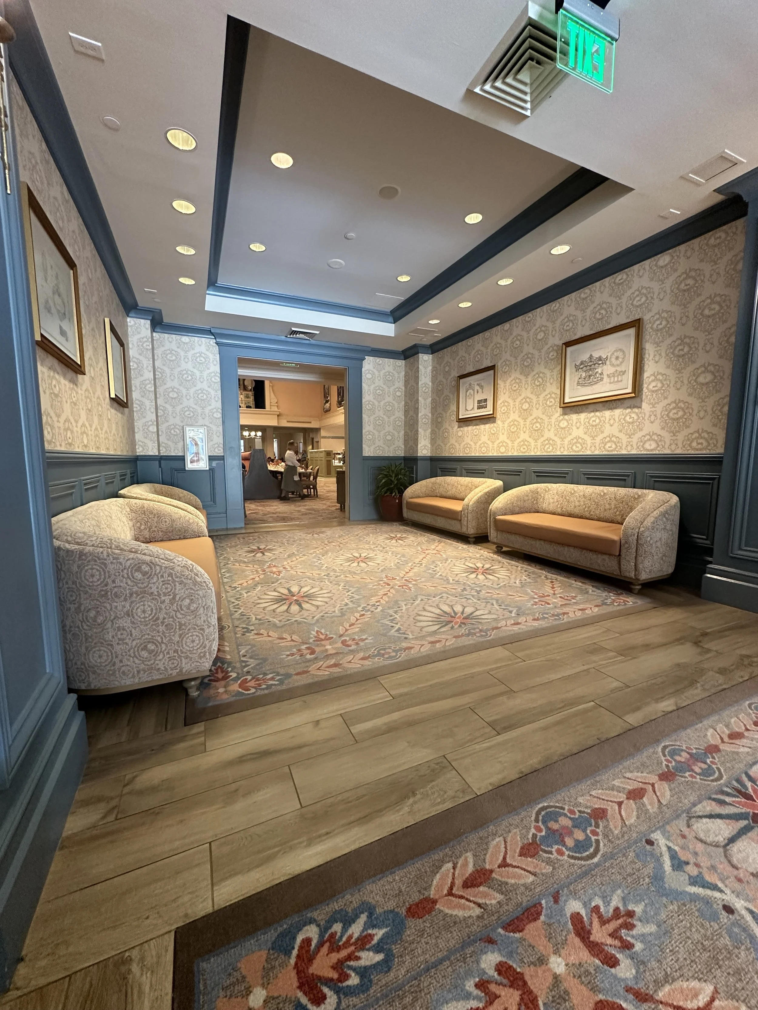 An elegant hotel lobby with patterned wallpaper, four beige sofas, a colorful area rug, and a doorway leading to another seating area.