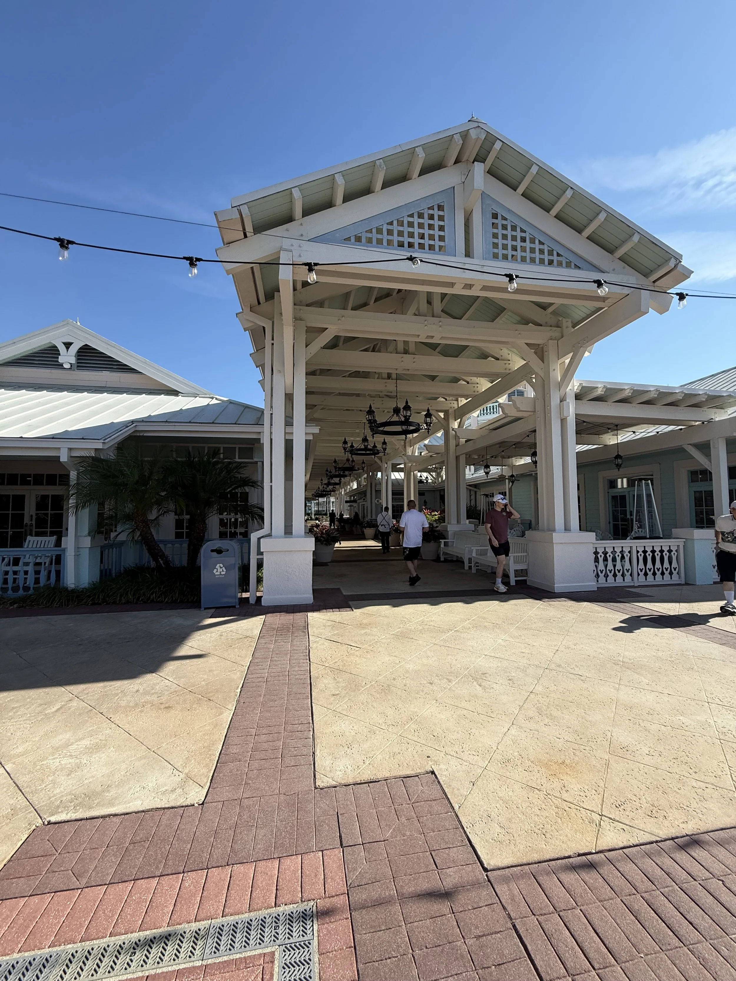 Outdoor shopping area with white wooden structures, hanging lights, a palm tree, and people walking under a clear blue sky.