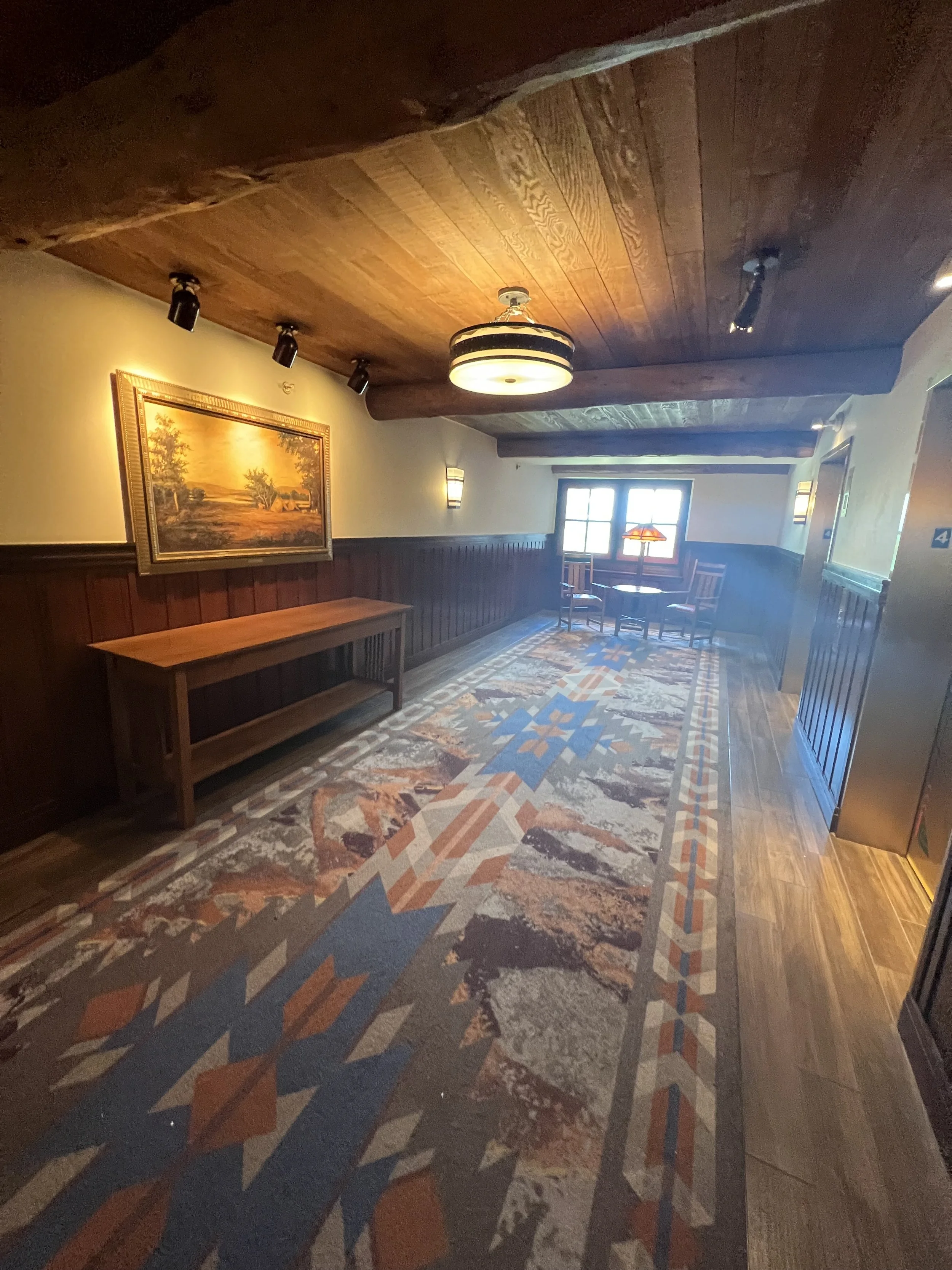 A cozy hallway area with wood paneling on the lower walls, an ornate landscape painting on the wall, a wooden bench, a patterned rug on the hardwood floor, and a small round table with chairs near a window, illuminated by warm lighting fixtures.