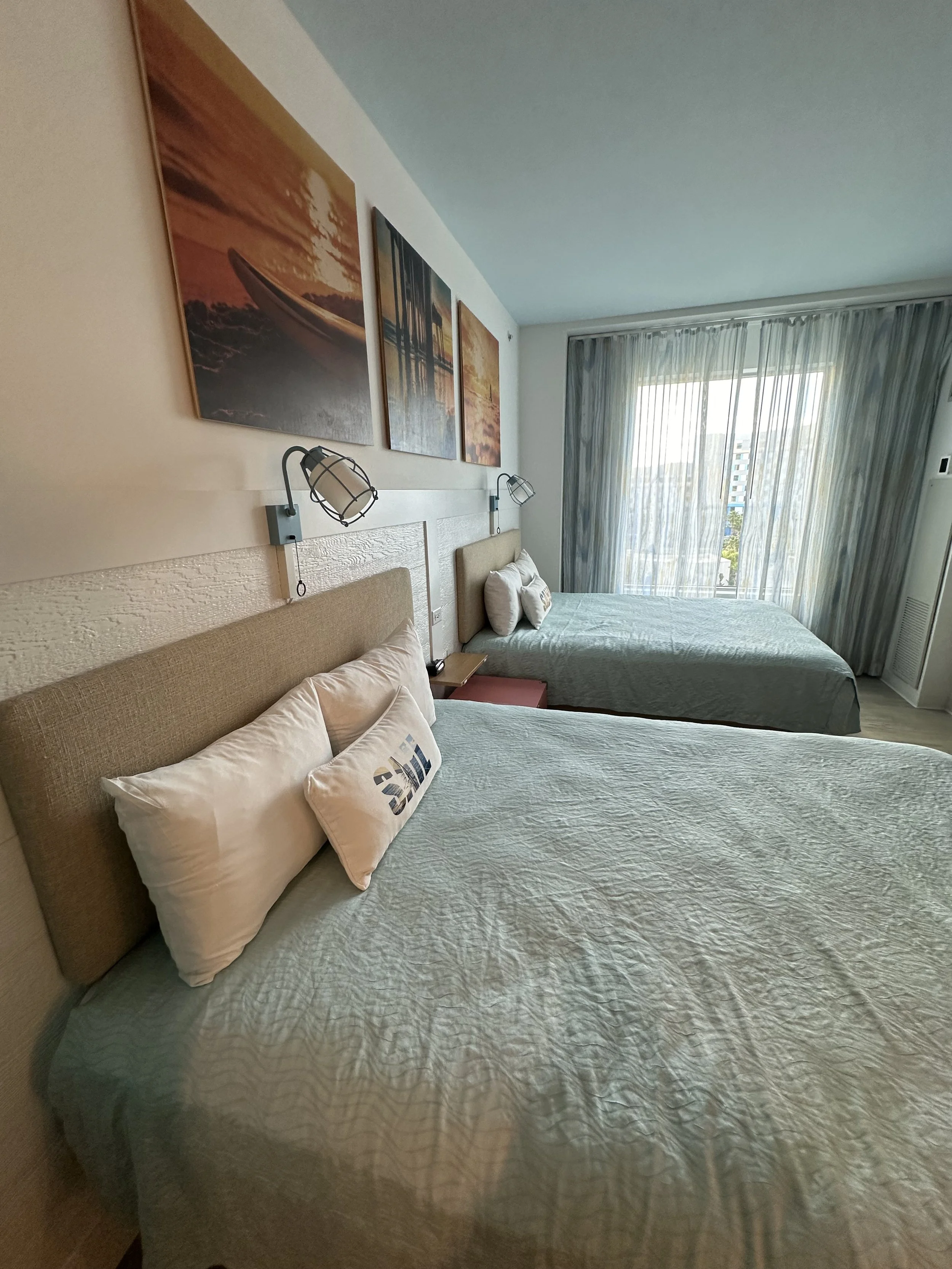 Hotel room with two beds, artwork of nature scenery above, and a large window with curtains