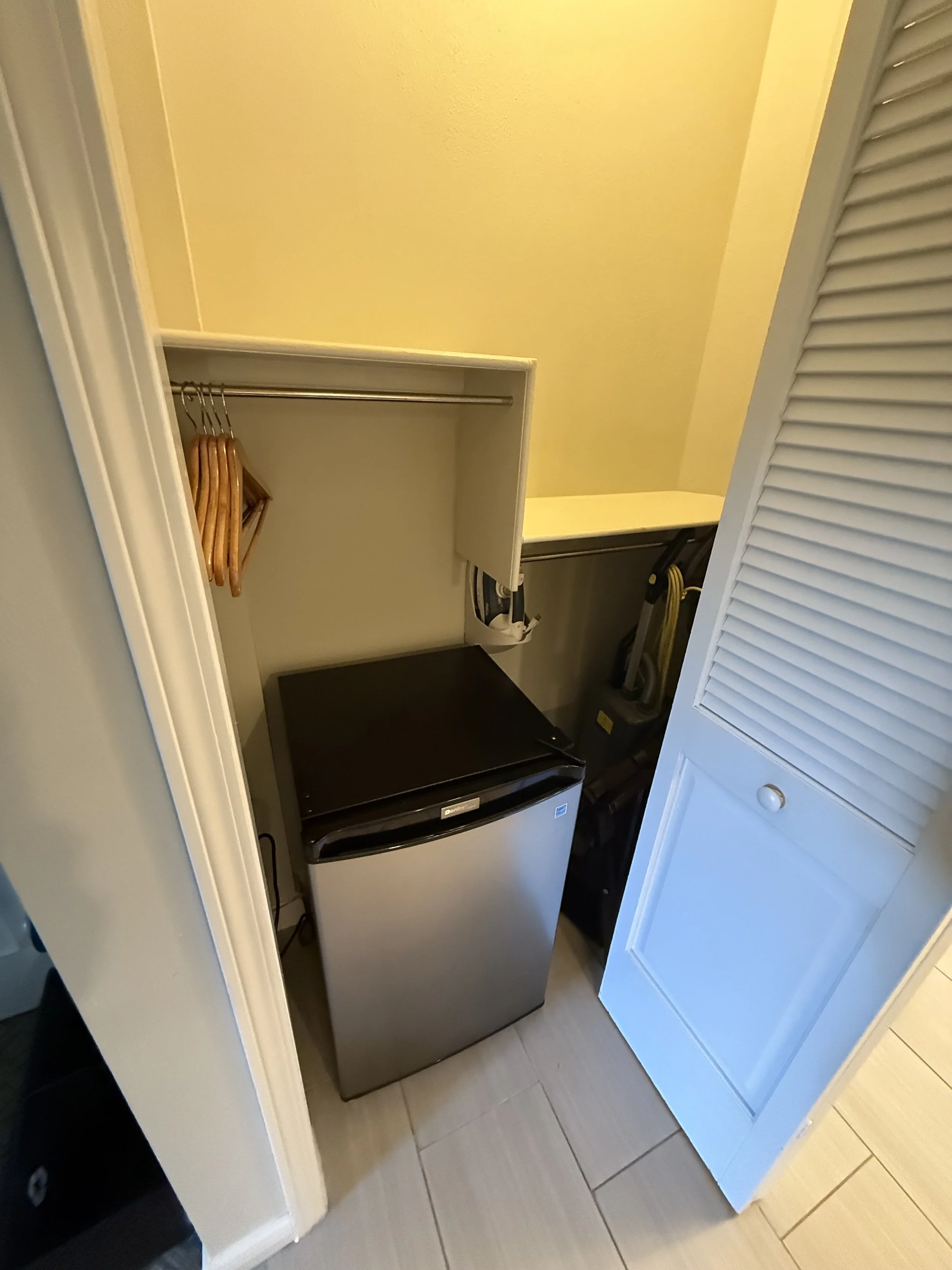 Small laundry closet with a mini refrigerator, a shelf with hangers, a vacuum cleaner, and a louvered door.