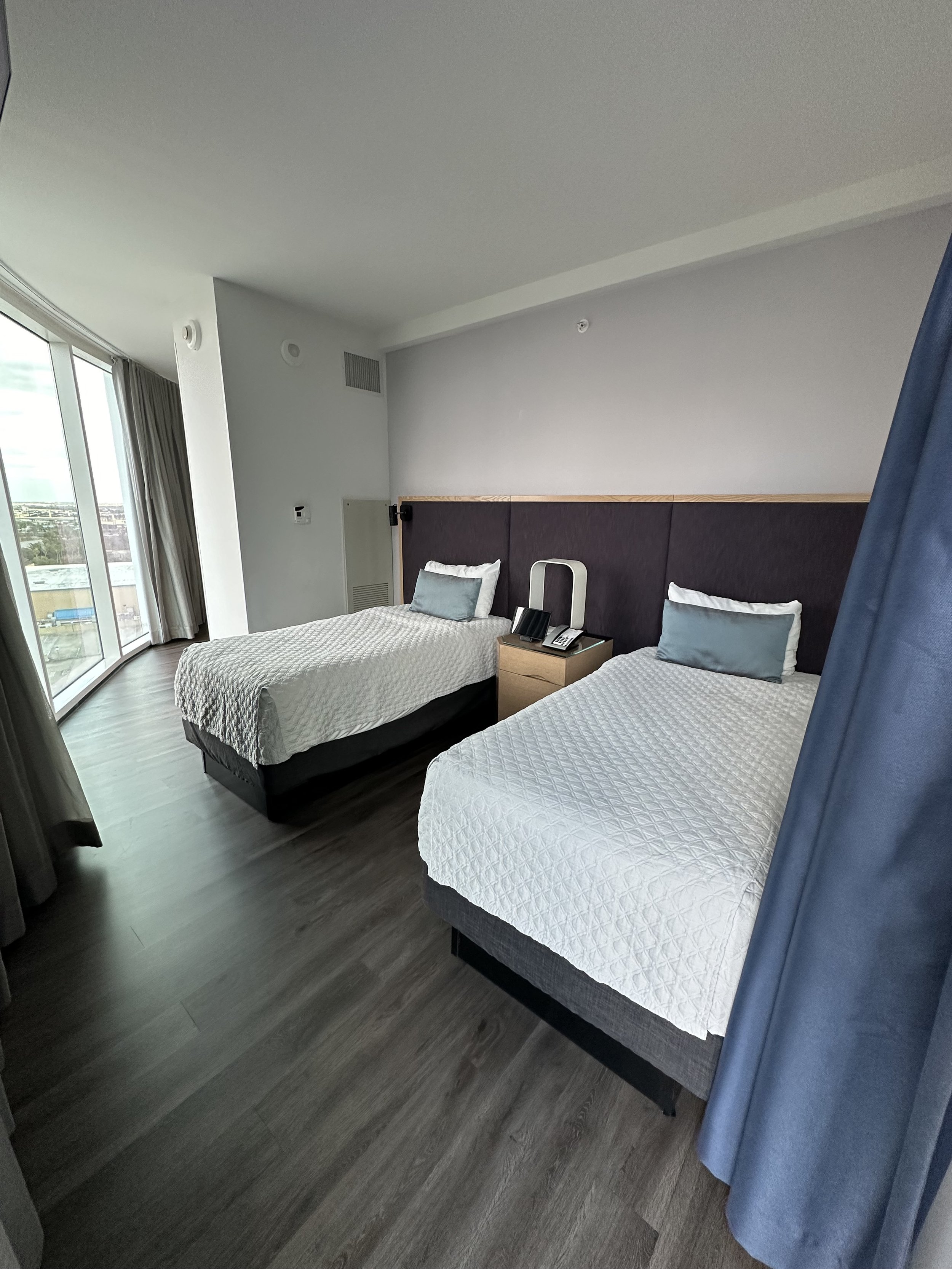 A hotel room with two single beds with white quilted bedding, each with a pillow and a decorative side pillow, a nightstand with a telephone between the beds, large window with blue curtains on the right, and glass sliding door on the left providing 