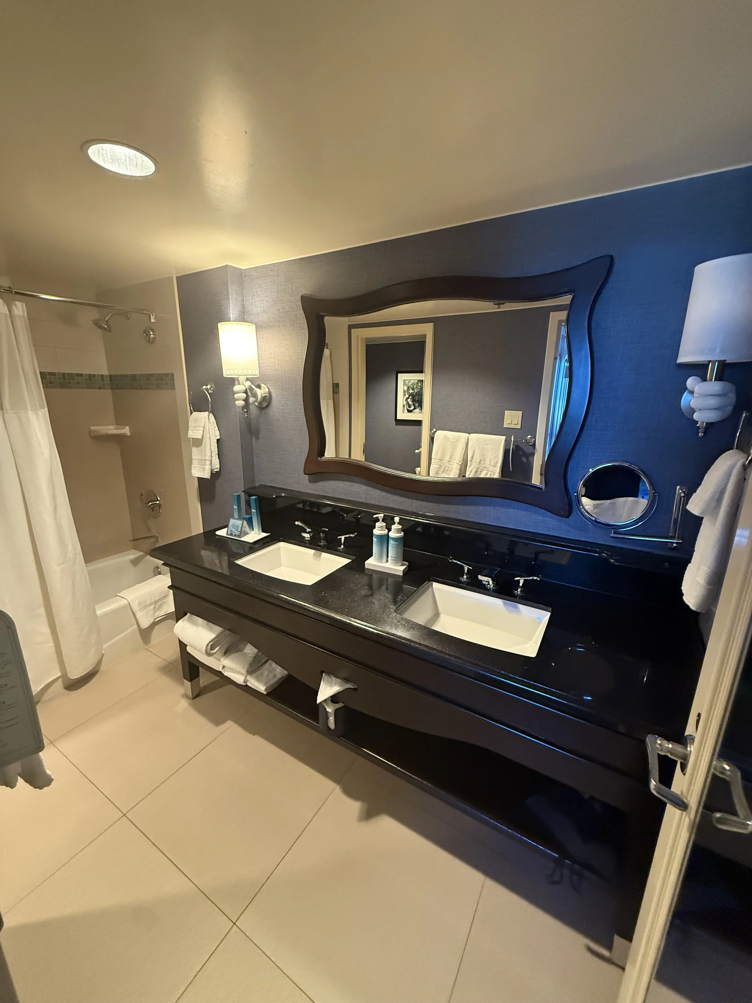 A hotel bathroom with a double sink vanity, large mirror, wall-mounted lamps, and a bathtub with a shower curtain on the left side.