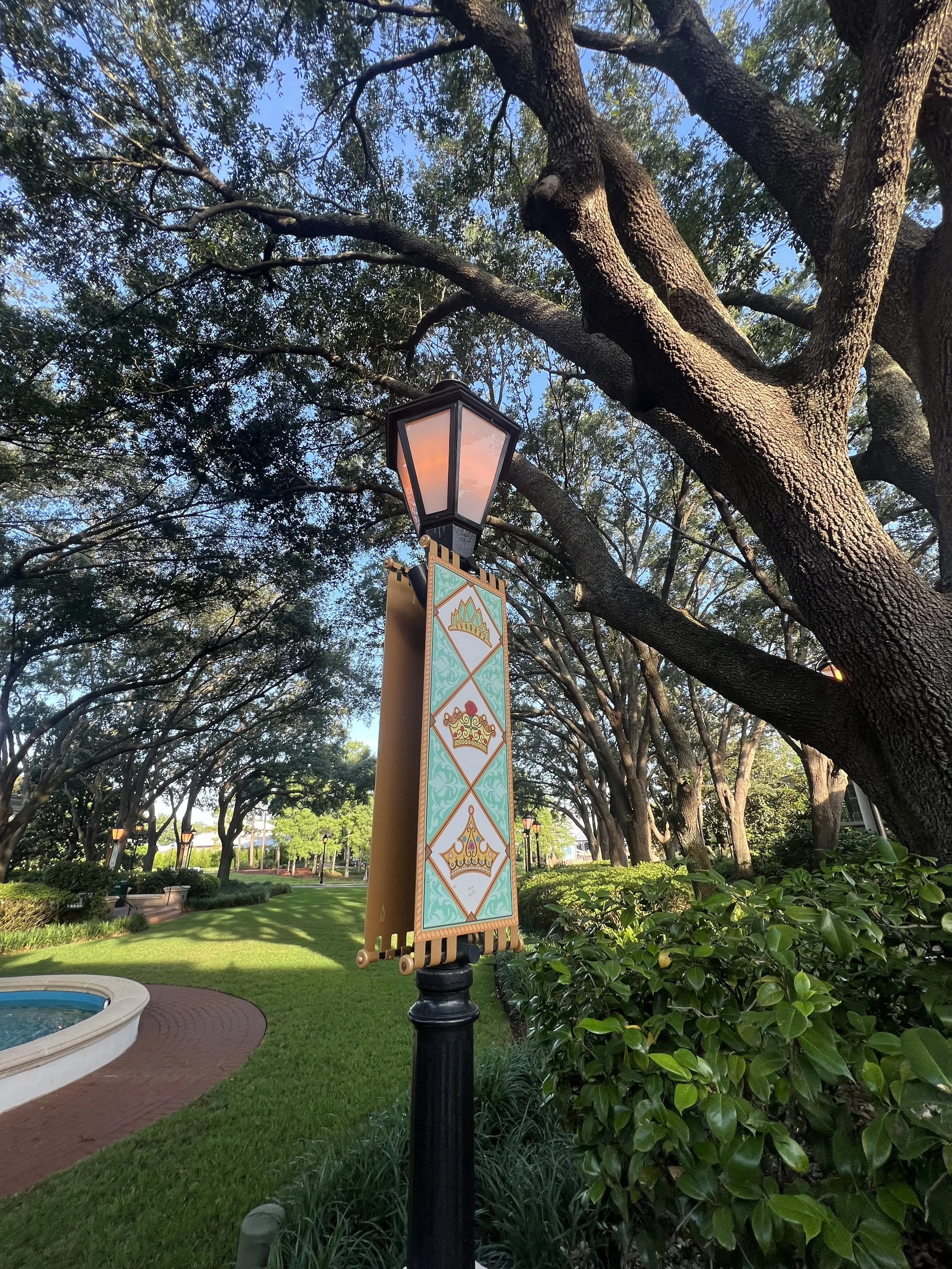 Decorative streetlamp with a mounting banner featuring colorful, ornate crowns in a park setting with lush green bushes, tall trees, and a paved pathway.