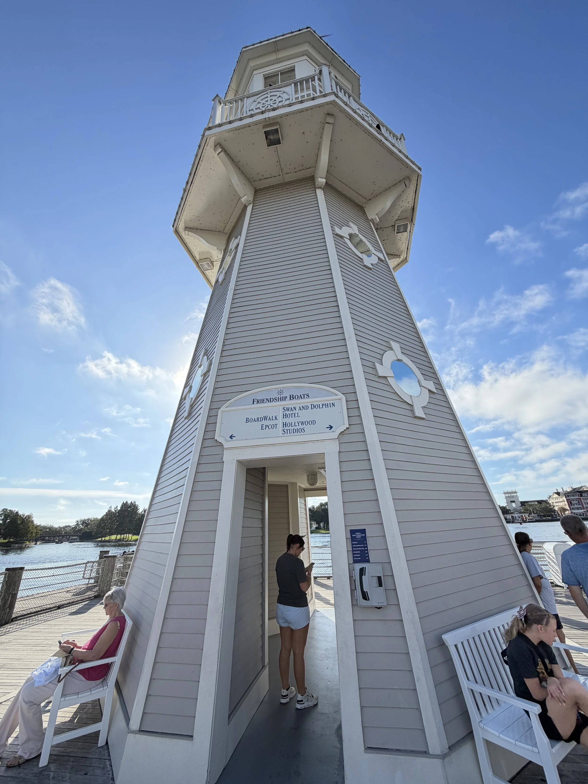 A lighthouse with a beige exterior, multiple windows, and a white balcony at the top, situated by a body of water with people sitting on benches and walking nearby.