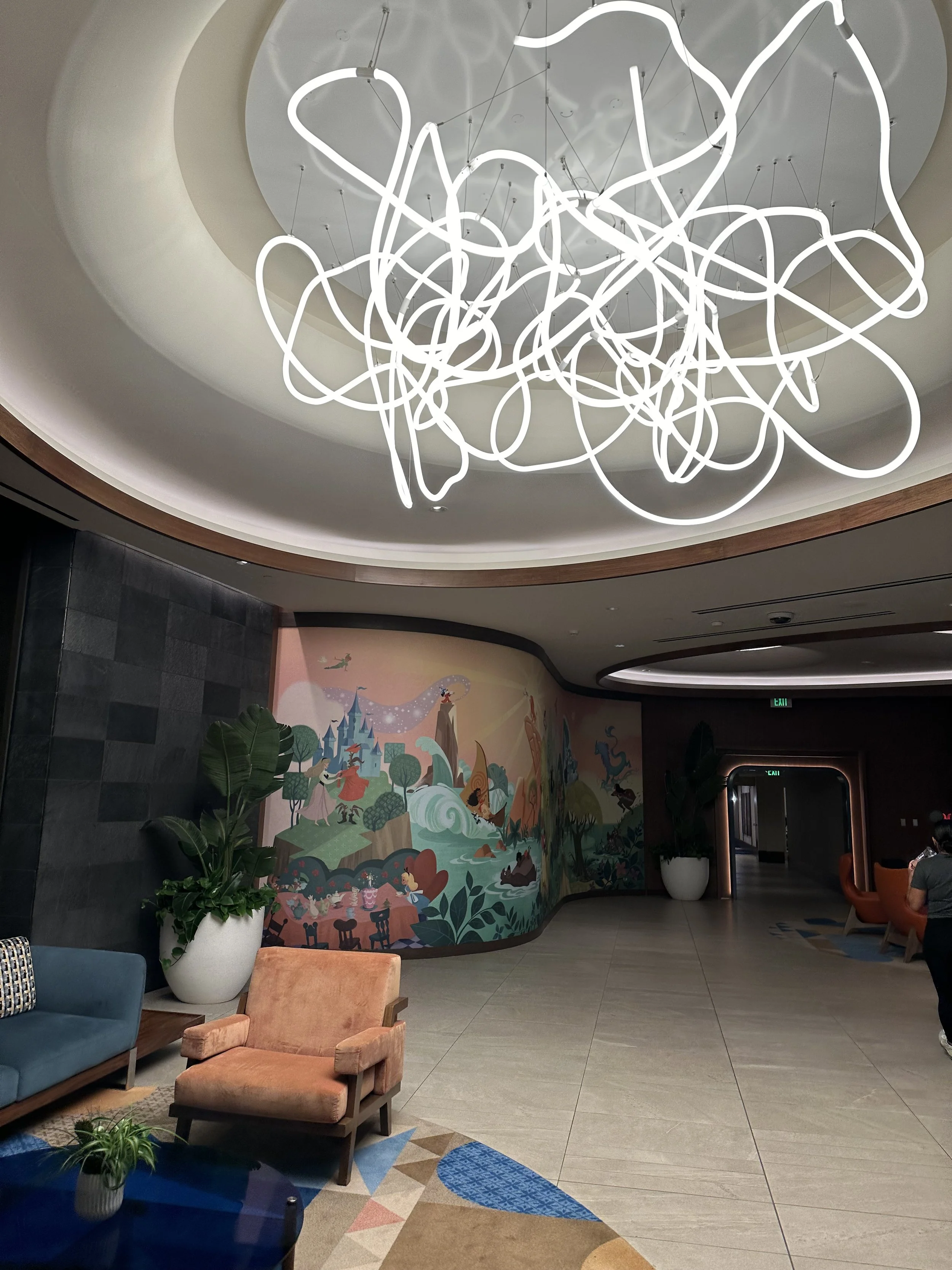 Interior view of a modern lobby with colorful mural on the wall, large potted plants, and contemporary furniture, including orange and blue chairs. A large abstract light fixture hangs from the ceiling.