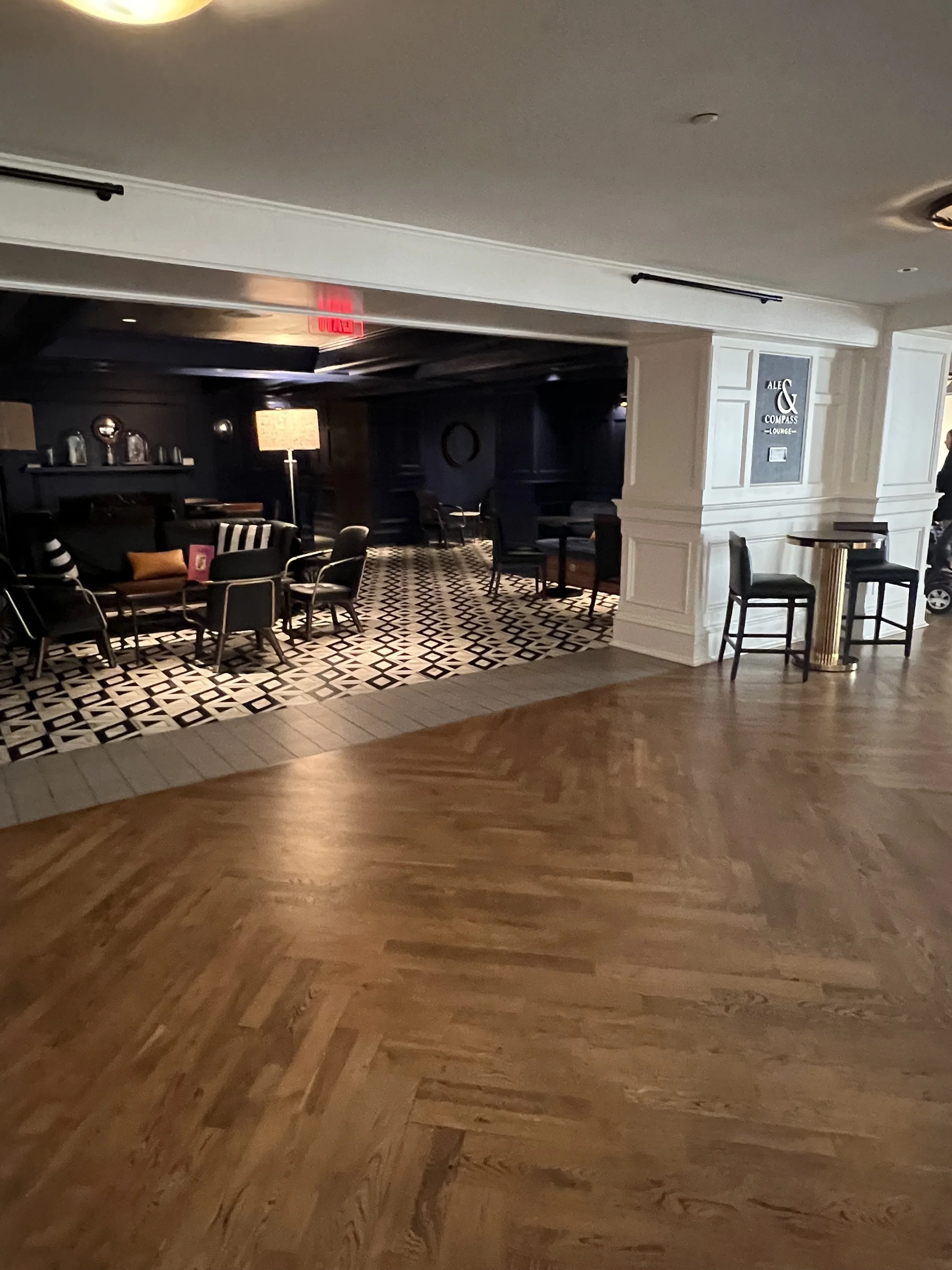 A hotel lobby with a mix of wooden and patterned tile flooring, a seating area with black chairs, a floor lamp, and a sign for Ale & Compass Lounge.