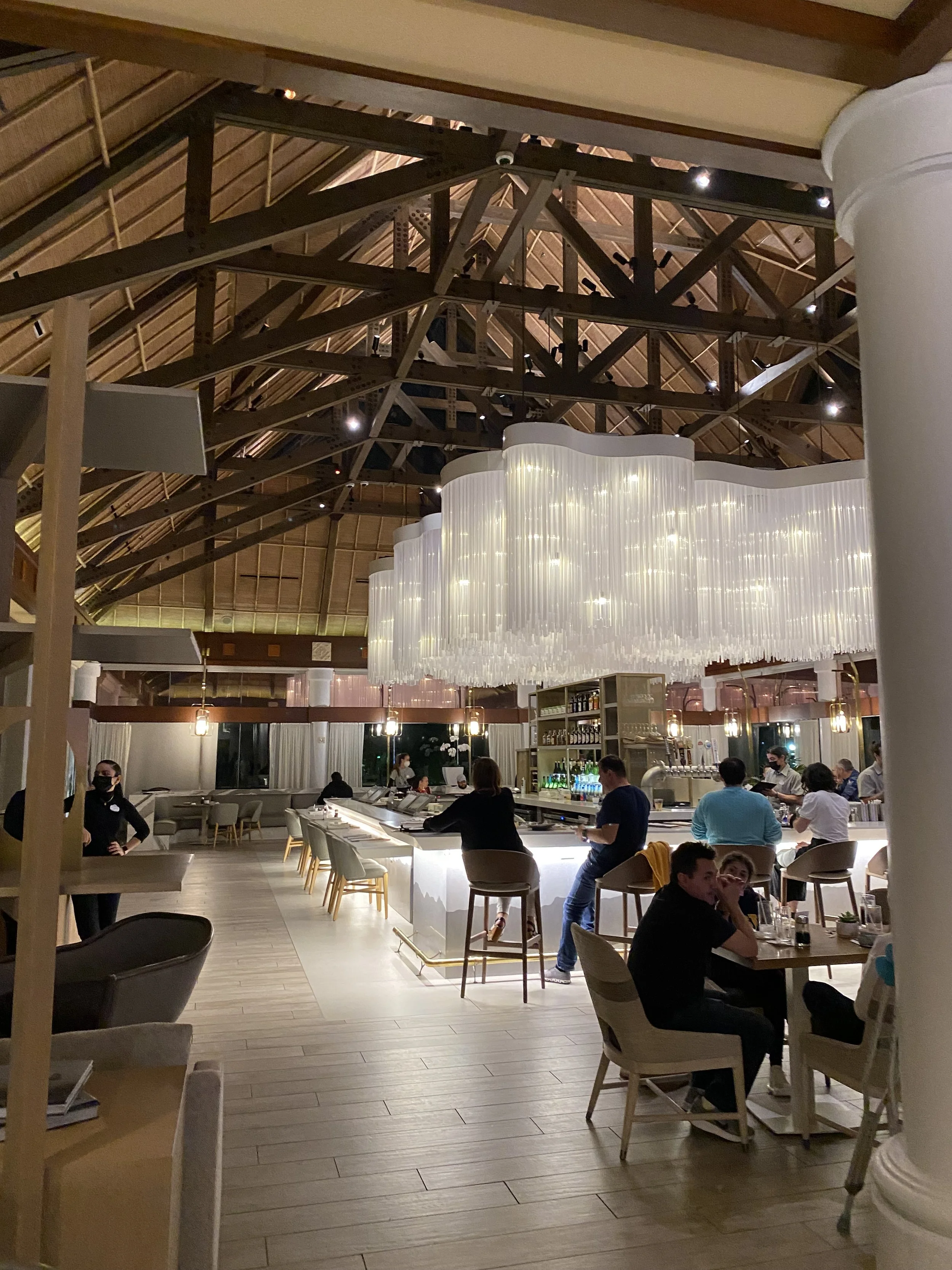Interior of a modern, upscale restaurant or bar with a high, wooden vaulted ceiling and large, white, wave-shaped chandelier. People are seated at the bar and tables, with some staff members attending to customers. The ambiance is warm and inviting w