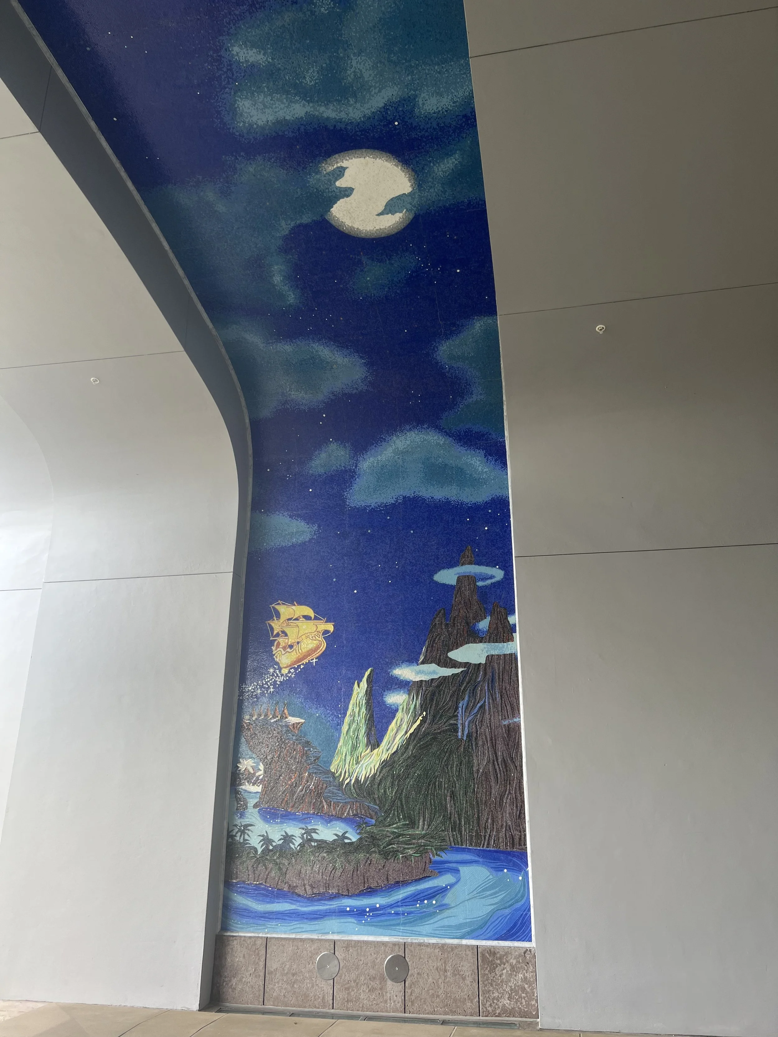 A mural of a fantasy landscape featuring mountains, a river, a sailing ship, clouds, stars, and a moon on an indoor wall.
