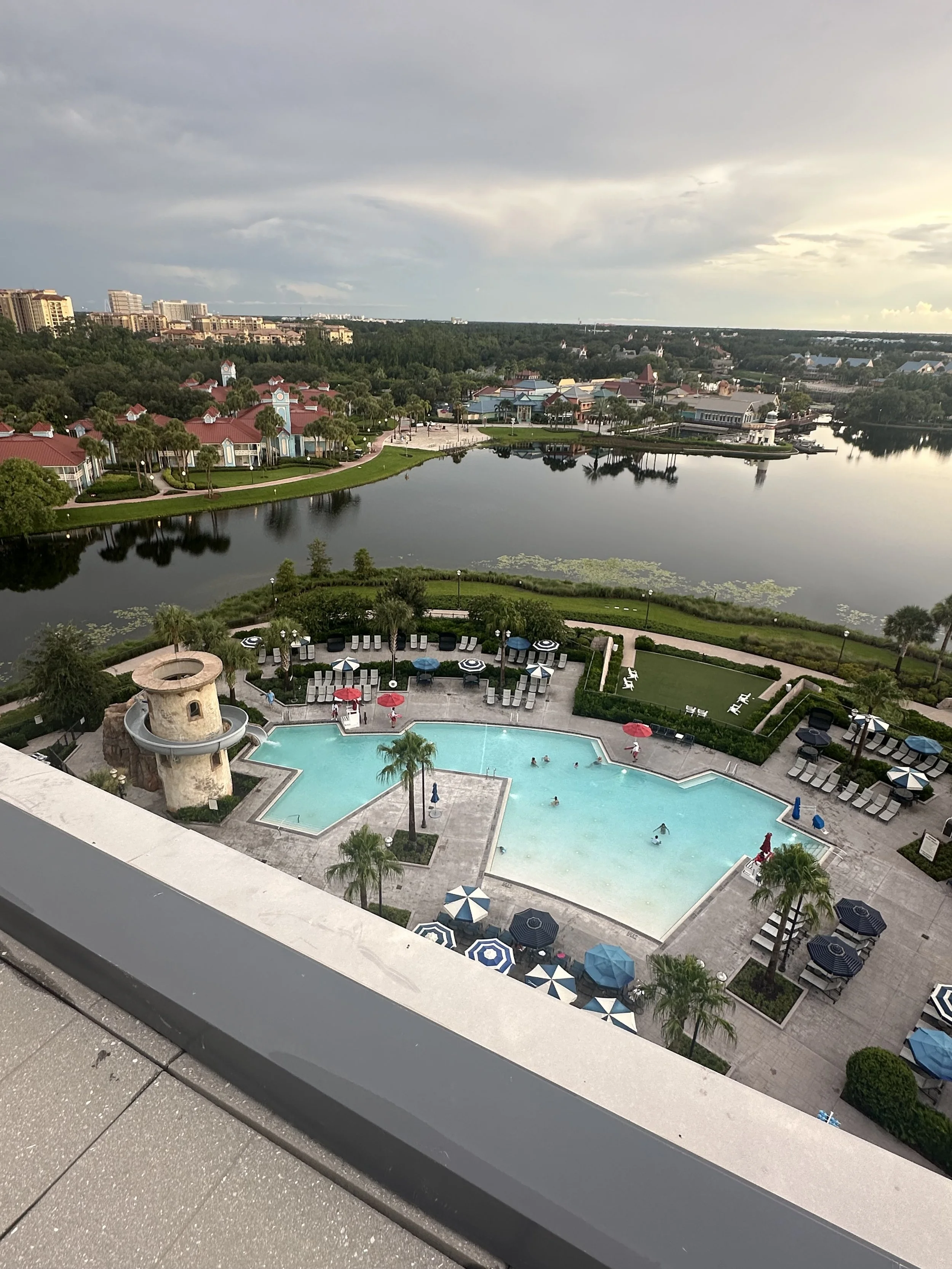 A rooftop view of a swimming pool area with lounge chairs, umbrellas, and a small tower, overlooking a pond with greenery and buildings in the background.