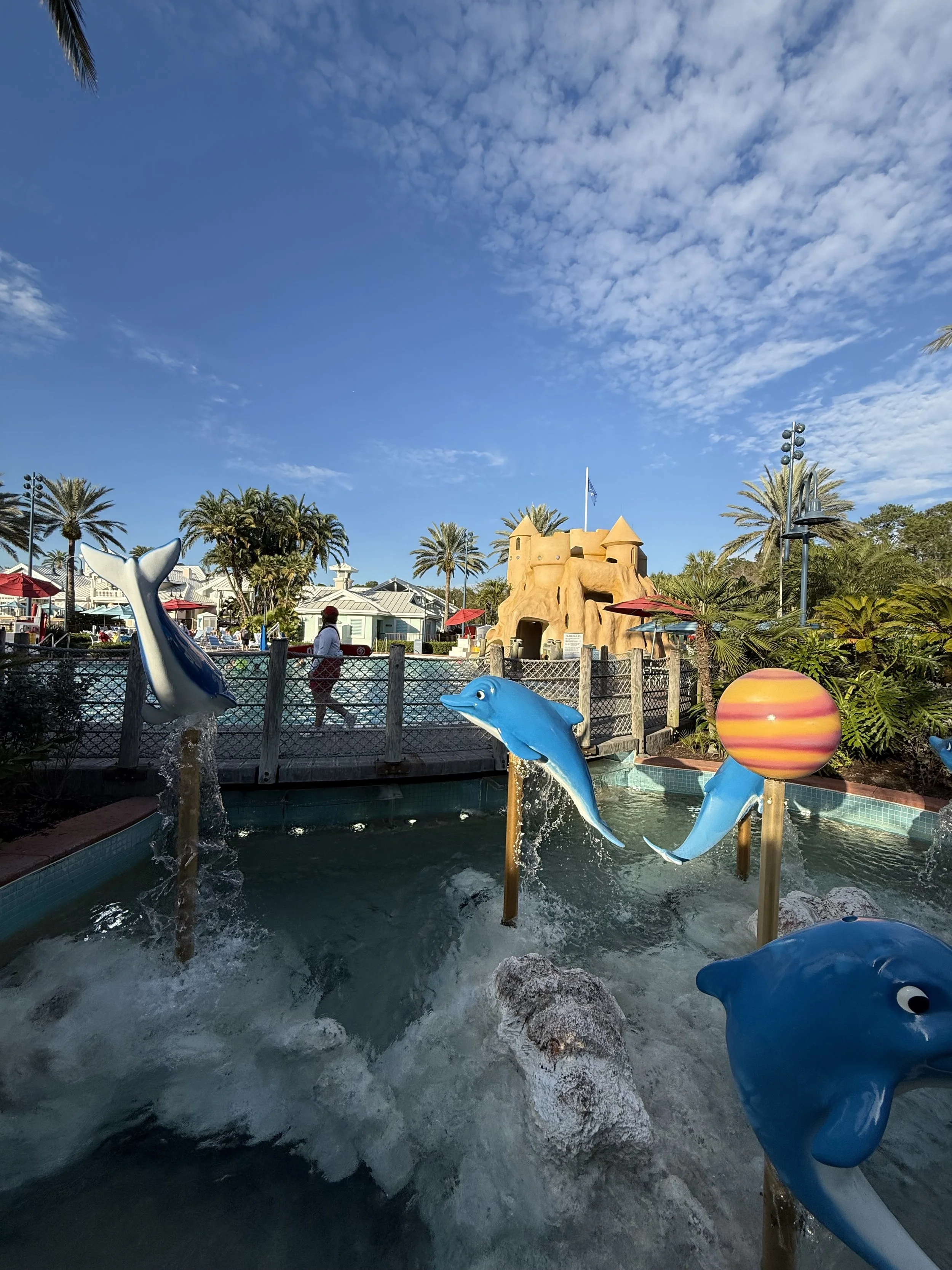 Colorful dolphin and ocean theme water fountain with a castle and palm trees in the background, part of a theme park or water park.