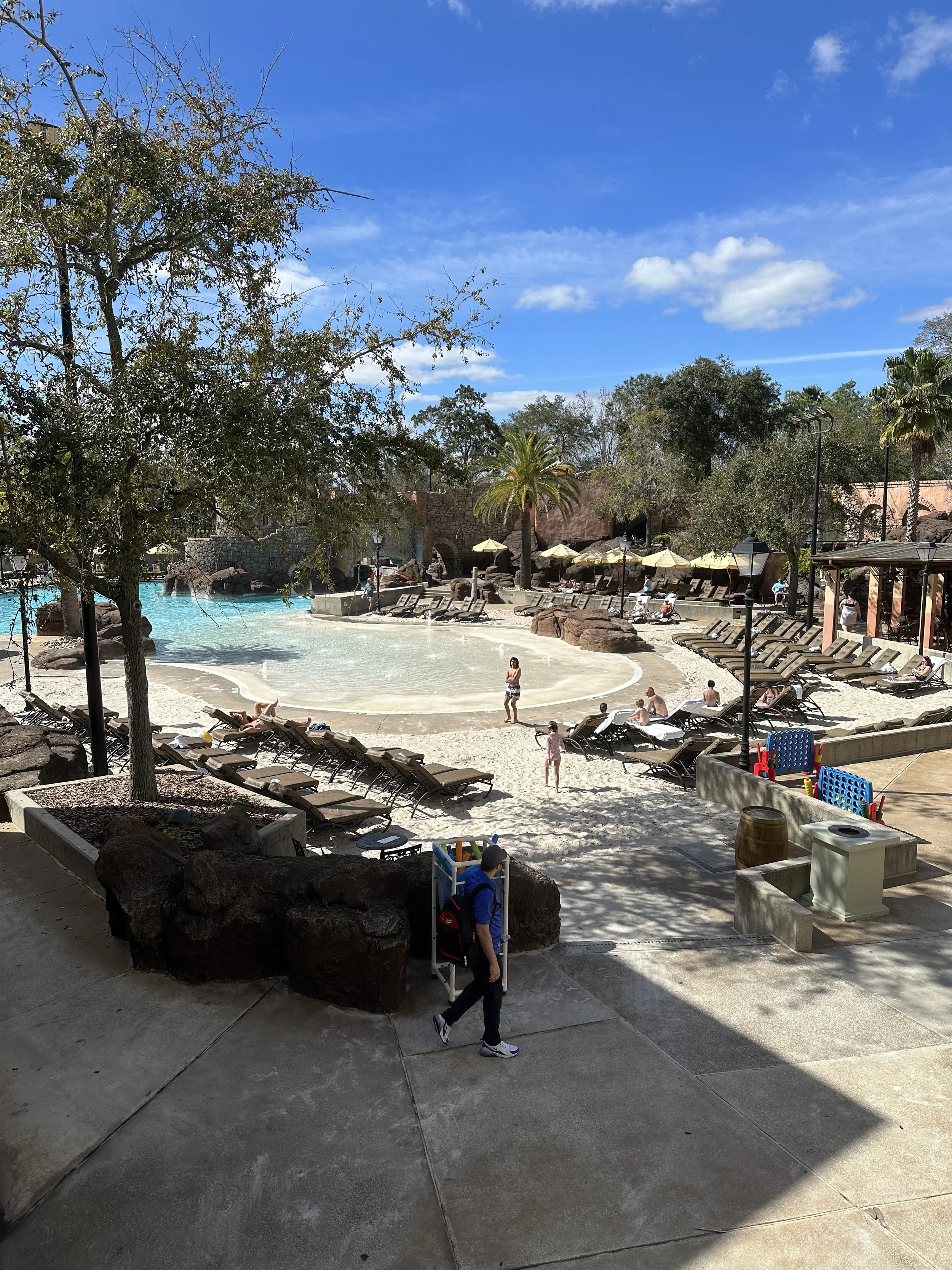A pool area at a resort or water park, with lounge chairs, umbrellas, trees, and a large pool shaped like a small lake. People are relaxing and swimming on a sunny day with a blue sky and some clouds.