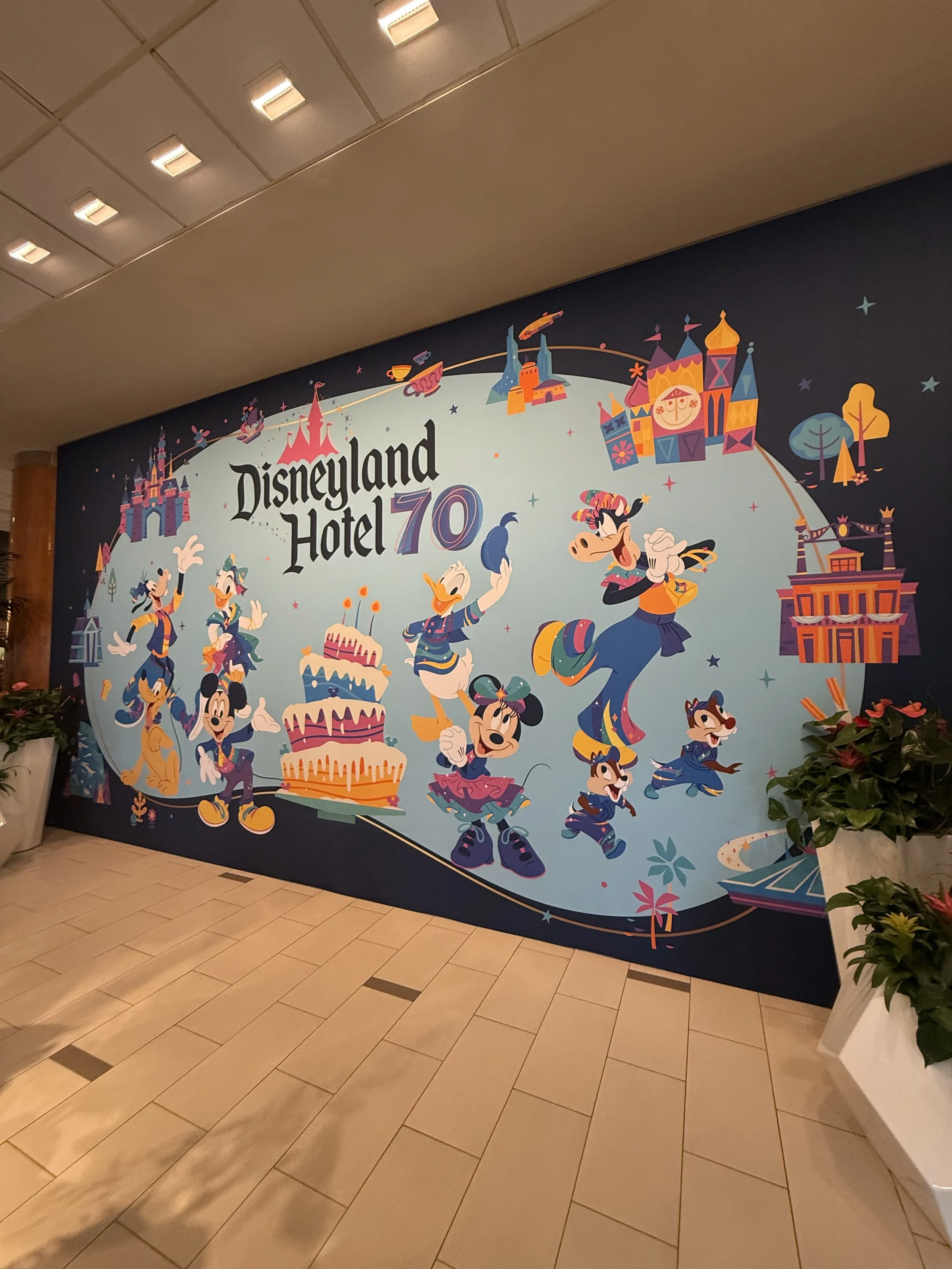 A celebration mural at Disneyland Hotel for its 70th anniversary, featuring Disney characters Mickey Mouse, Minnie Mouse, Donald Duck, and others, with Disney castle and themed illustrations.