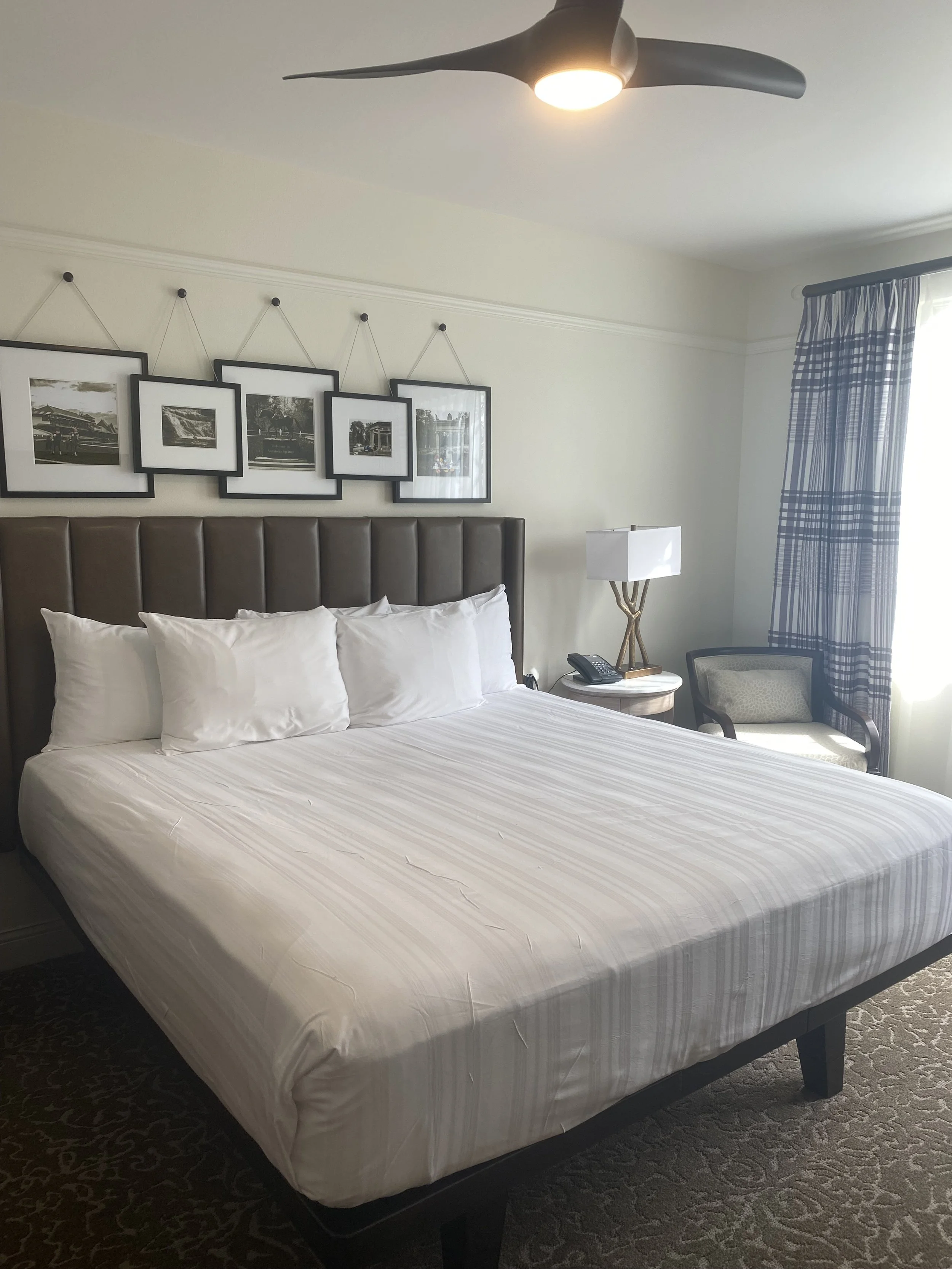 A hotel room with a large bed, six white pillows, a brown headboard, a nightstand with a lamp and phone, a chair next to a window with blue and white curtains, and black and white framed pictures on the wall. A ceiling fan light is on.