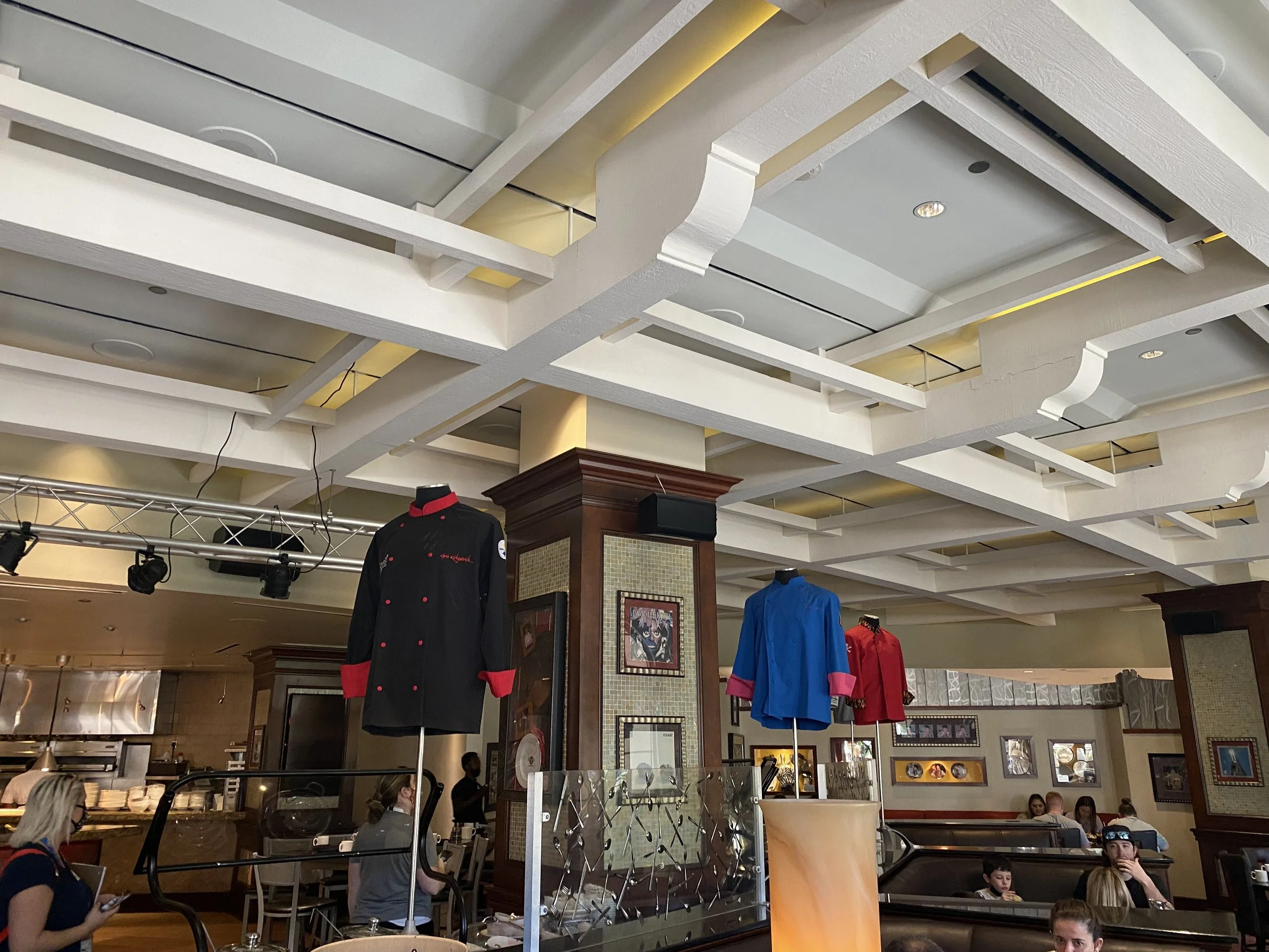 Interior of a restaurant with chefs' jackets in red, black, and blue hanging from display stands, and customers dining in the background.