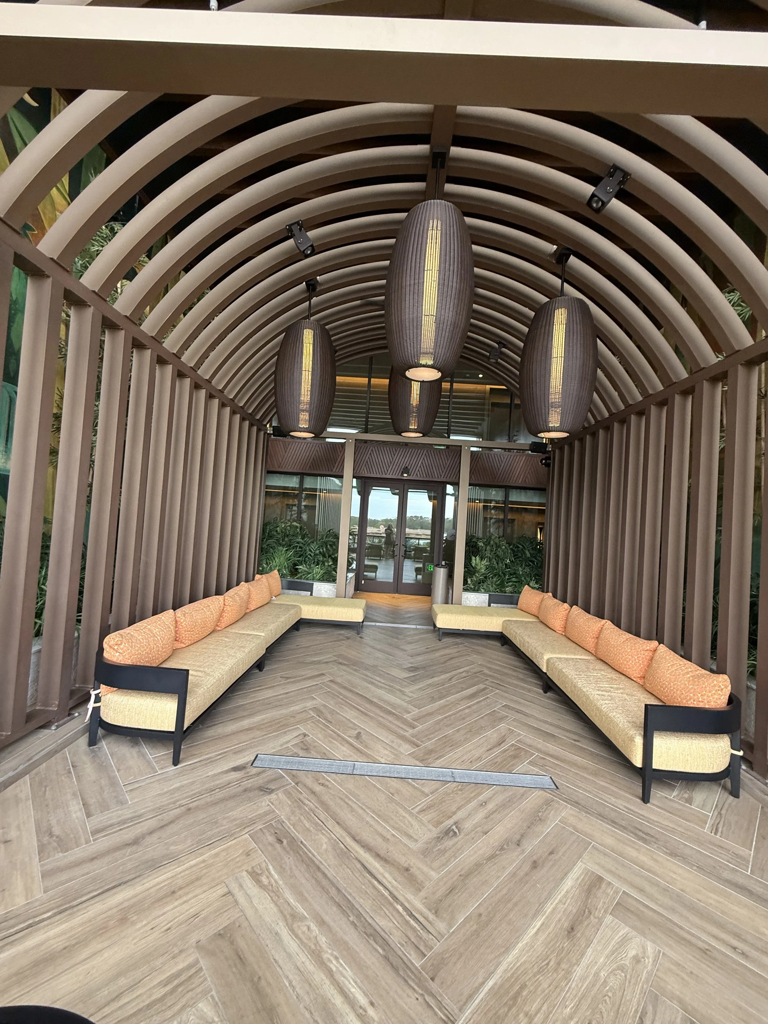 Indoor seating area with two long beige couches along each side, orange cushions, wooden flooring, and hanging pendant lights in a curved wooden framework ceiling.