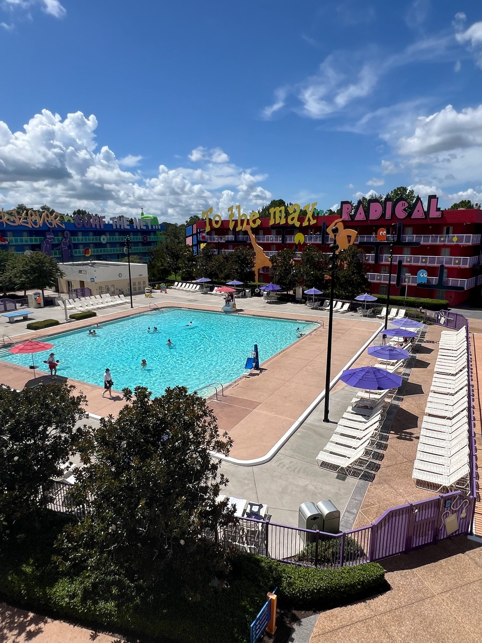 A colorful outdoor pool with people swimming, surrounded by lounge chairs and umbrellas, with vibrant buildings and signs in the background.