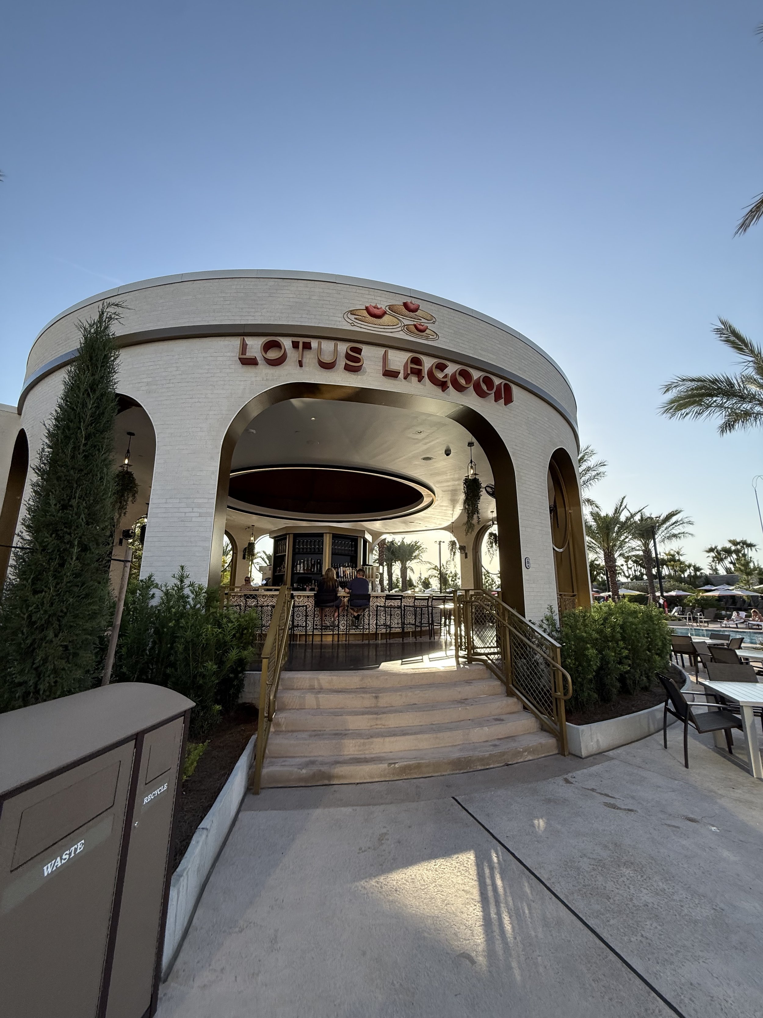 Exterior view of Lotus Lagoon, a bar or restaurant with a curved entrance, outdoor seating, palm trees, and a clear blue sky.