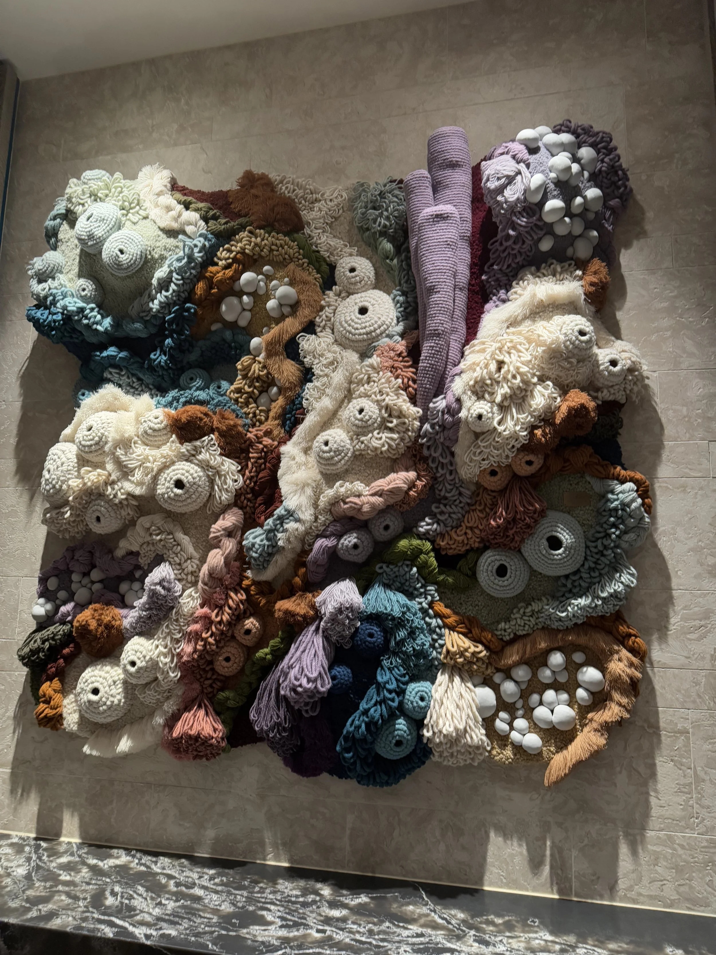 Colorful textured wall art made of various knitted and crocheted yarn shapes and clusters, including circular and elongated forms, arranged in a mixed, organic composition on a beige tiled wall.