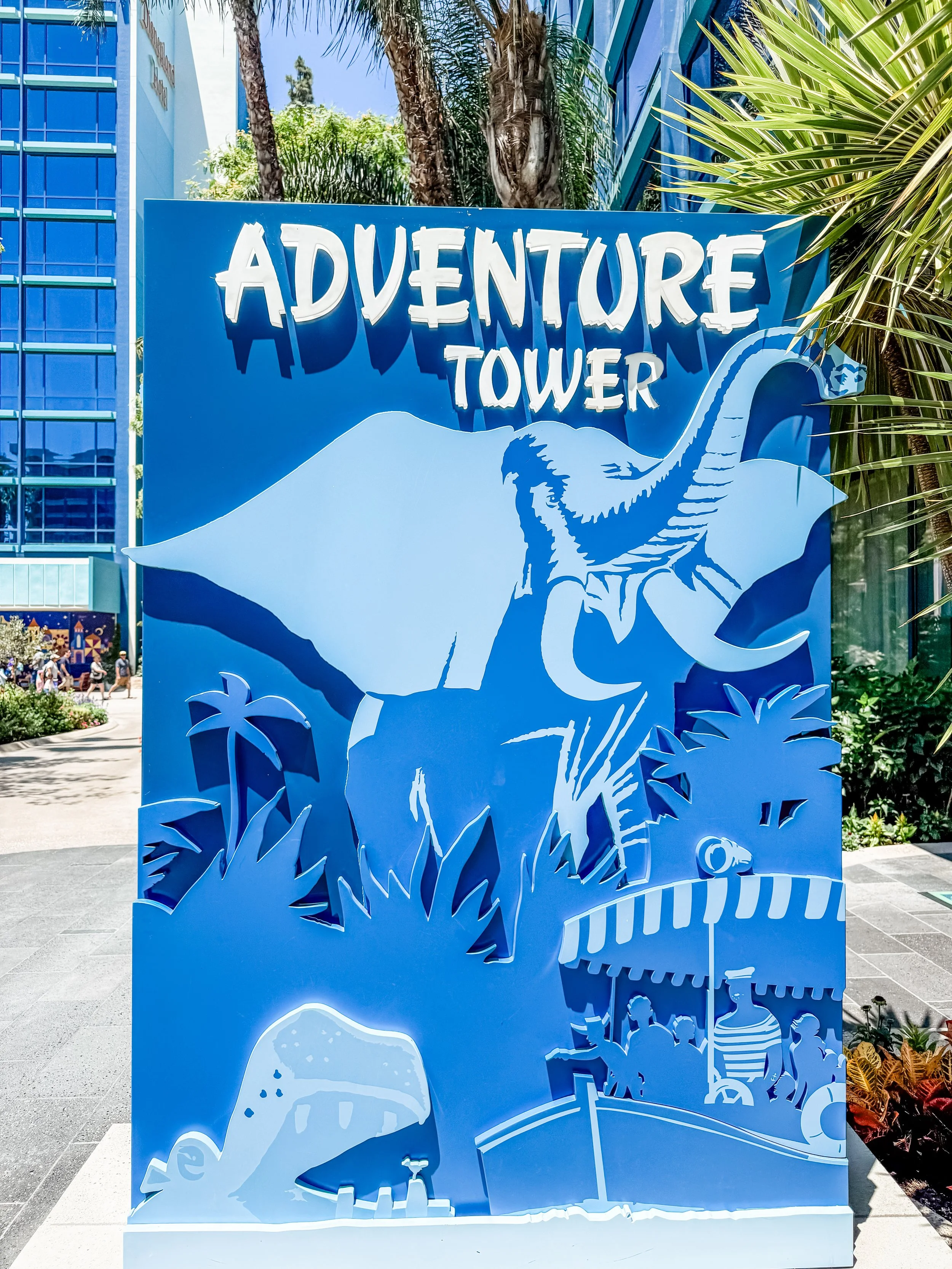 Blue sign reading 'Adventure Tower' with a cutout of an elephant, trees, and a boat with people underneath an awning.