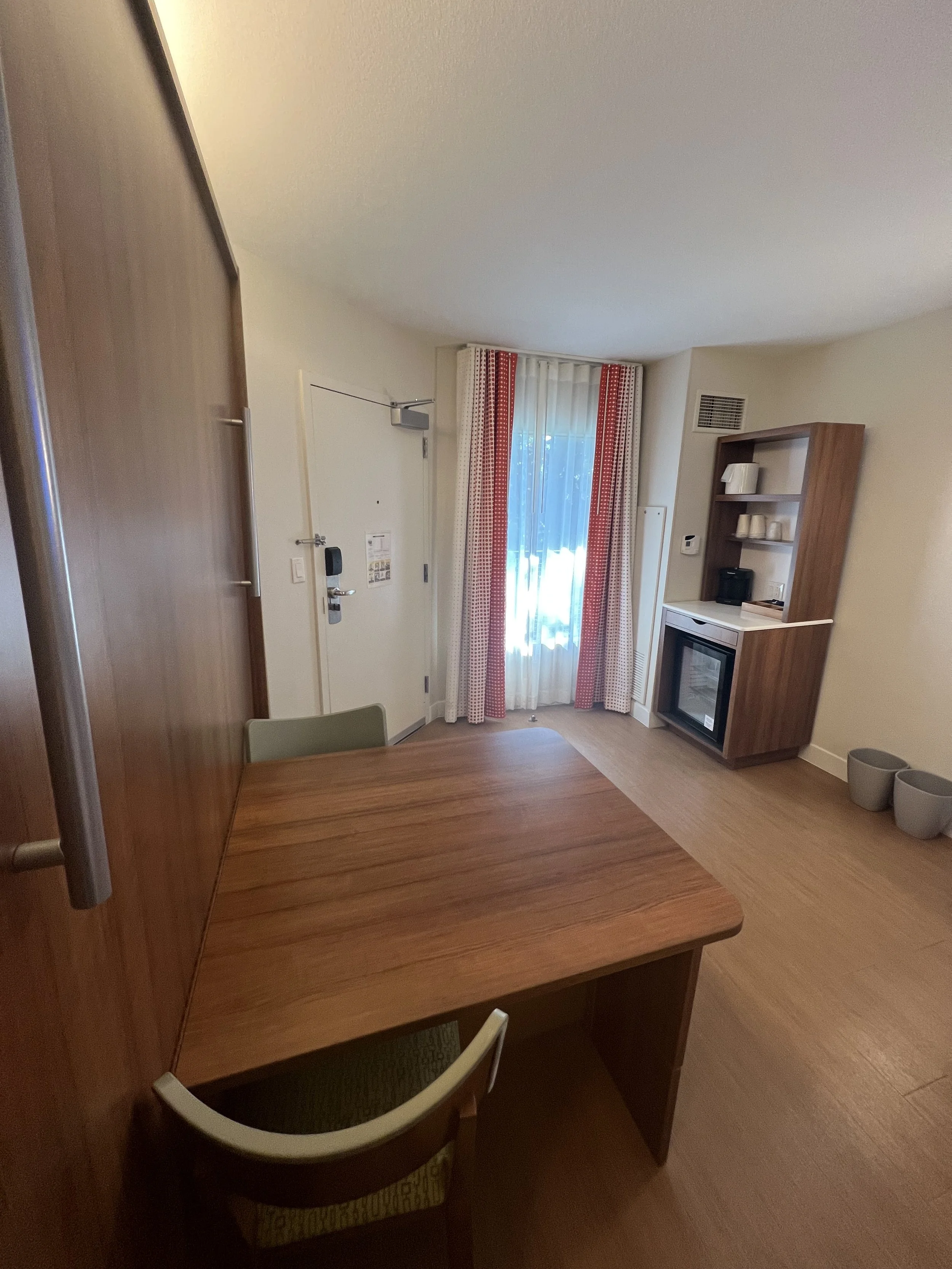 Hotel hotelroom with wooden table, chairs, curtains, a small built-in shelving unit with dishes, a mini fridge, and a door leading outside.