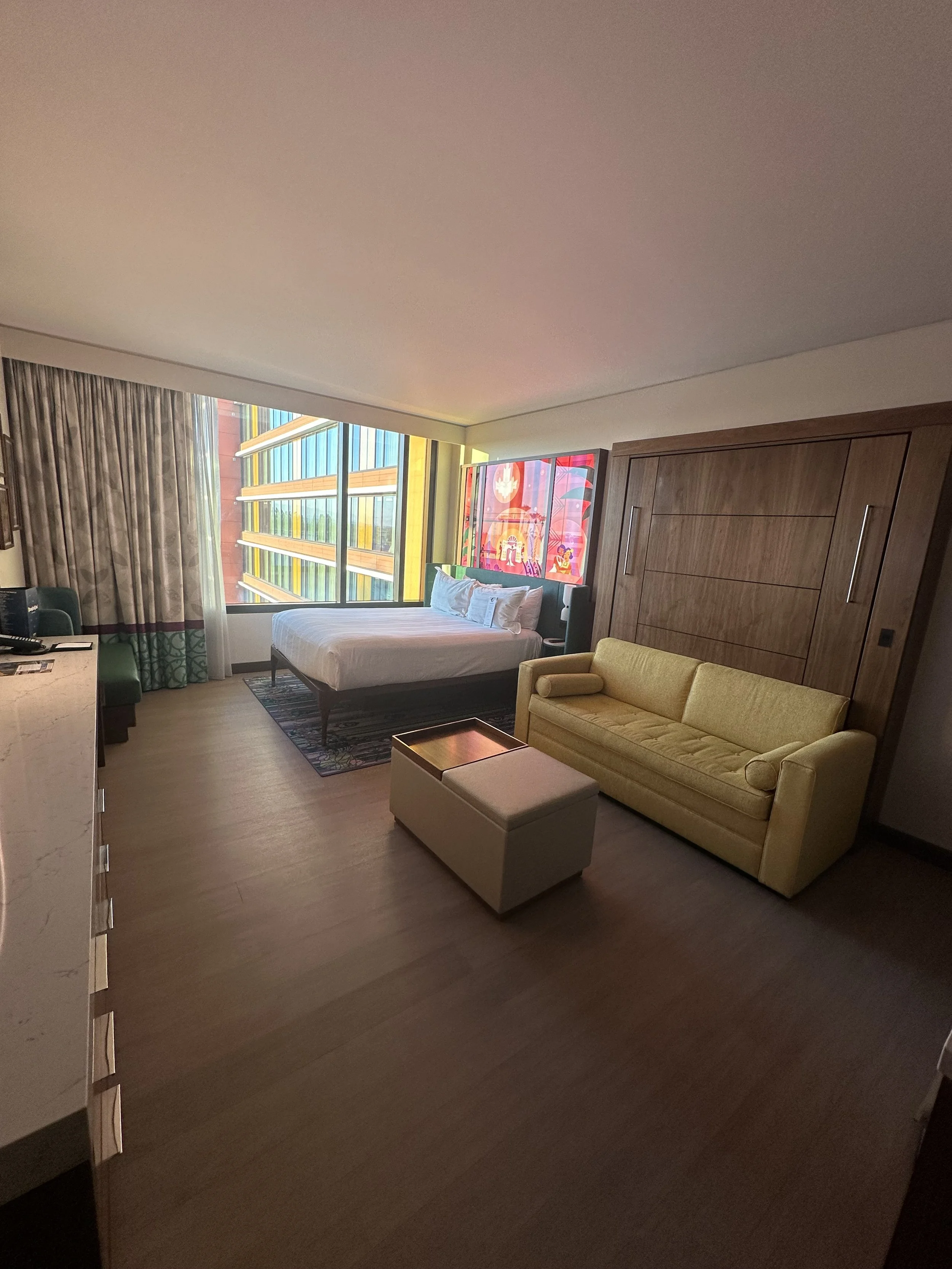 Hotel room with a bed near large windows, a yellow sofa, an ottoman, and a wooden wall.