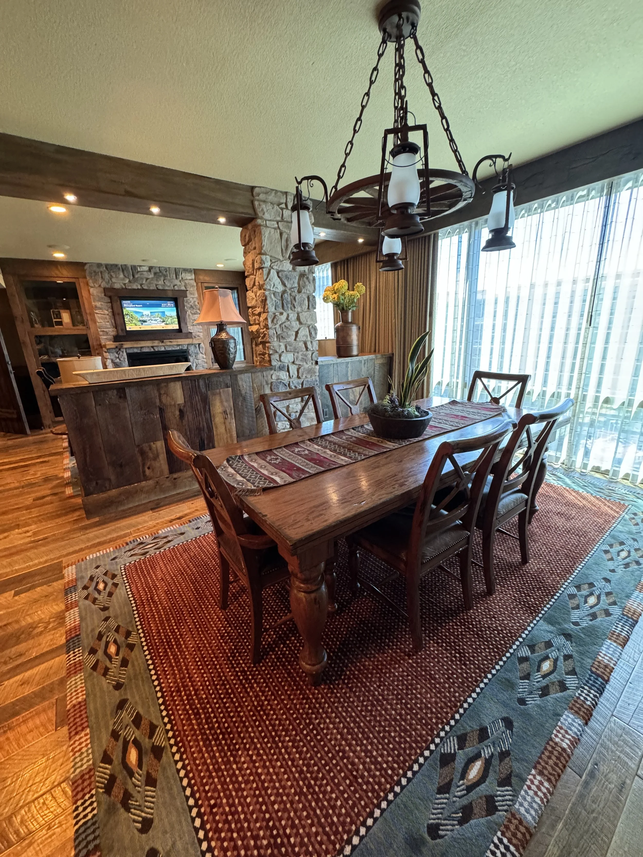Wooden dining table with six chairs, a decorative rug underneath, and a centerpiece plant. Overhead chandelier with lantern-style lights. Sunlight through large windows with vertical blinds.