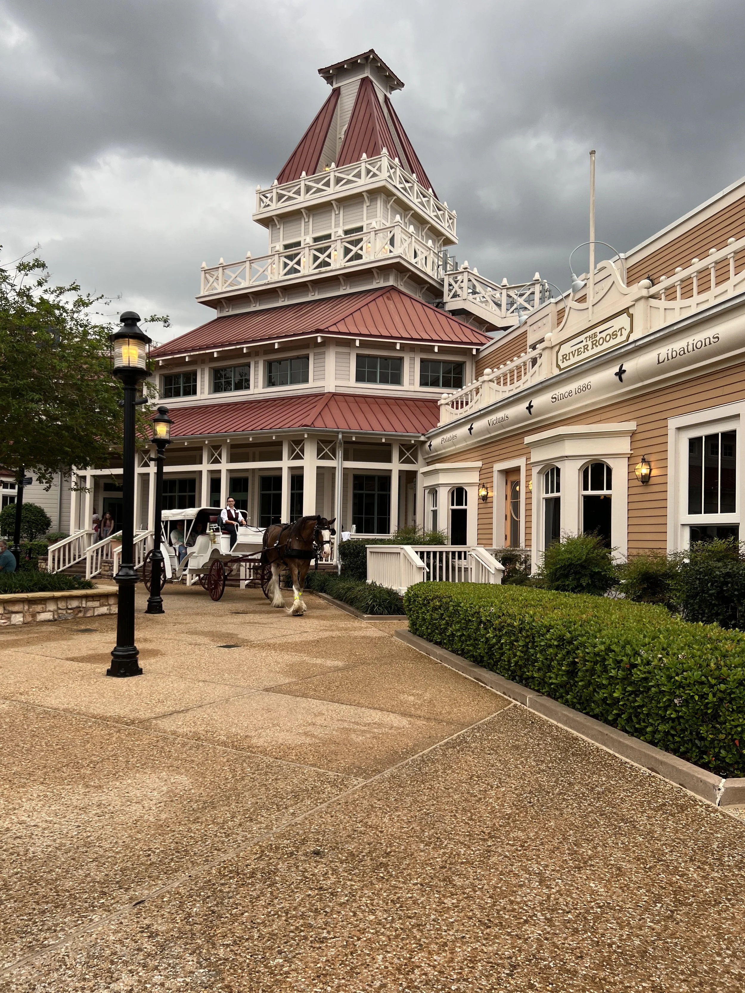 A building with a red, multi-tiered roof, white trim, surrounded by a walkway, shrubs, and trees. A horse-drawn carriage is in front of the building, and the sky above is overcast.