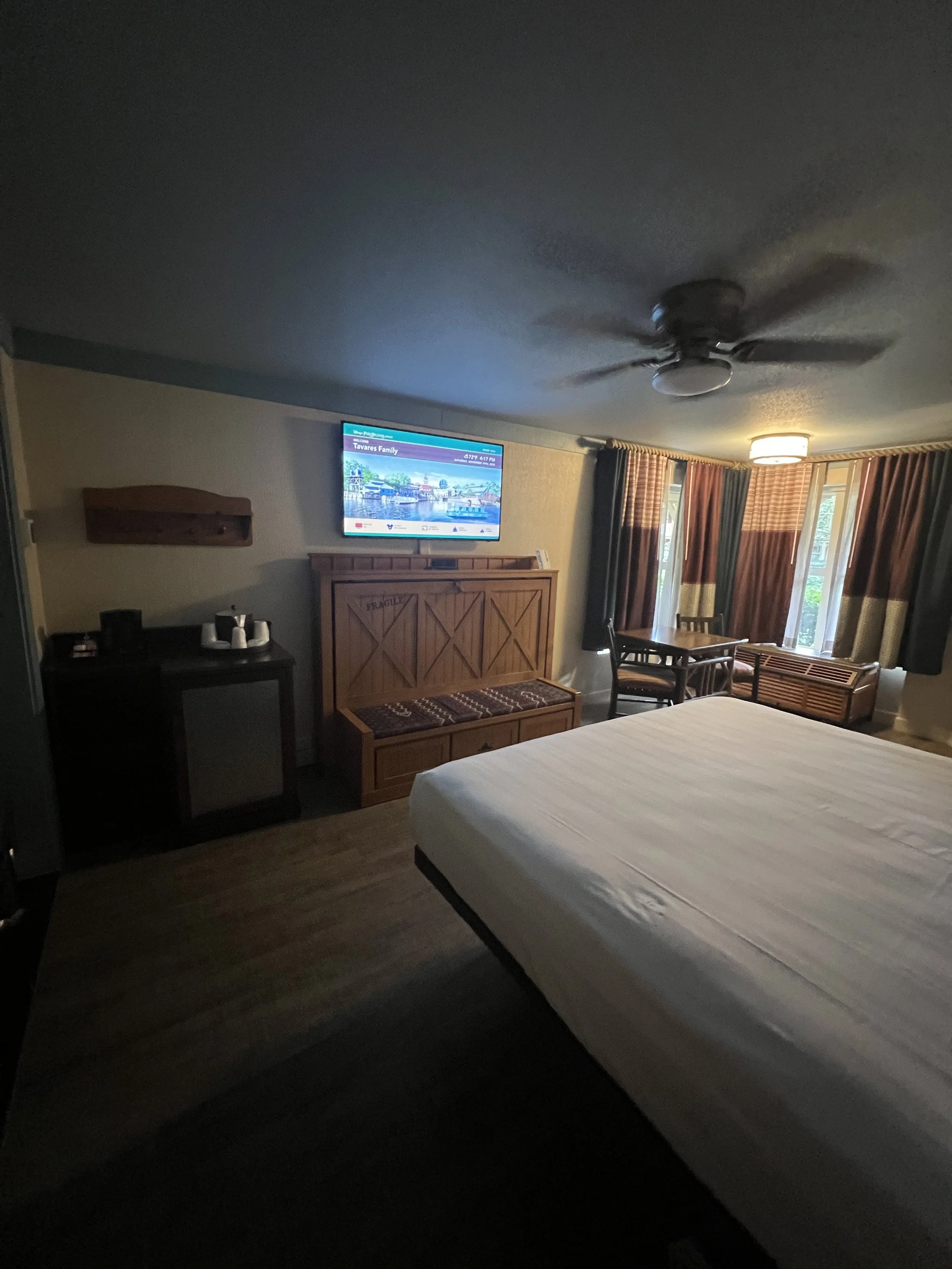 Hotel room with a bed, a wooden cabinet with a TV mounted above, a small table with chairs near the window, and curtains.