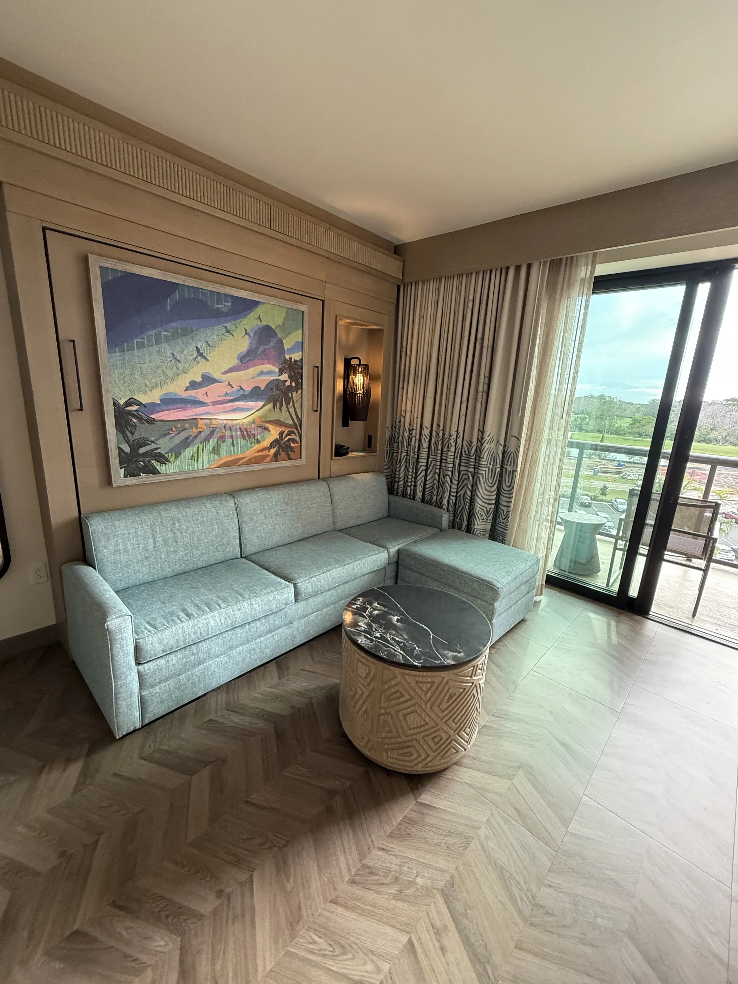 Living room with a light grey sectional sofa, a small round marble-topped coffee table, artwork on the wall, and sliding glass door leading to a balcony with outdoor furniture.