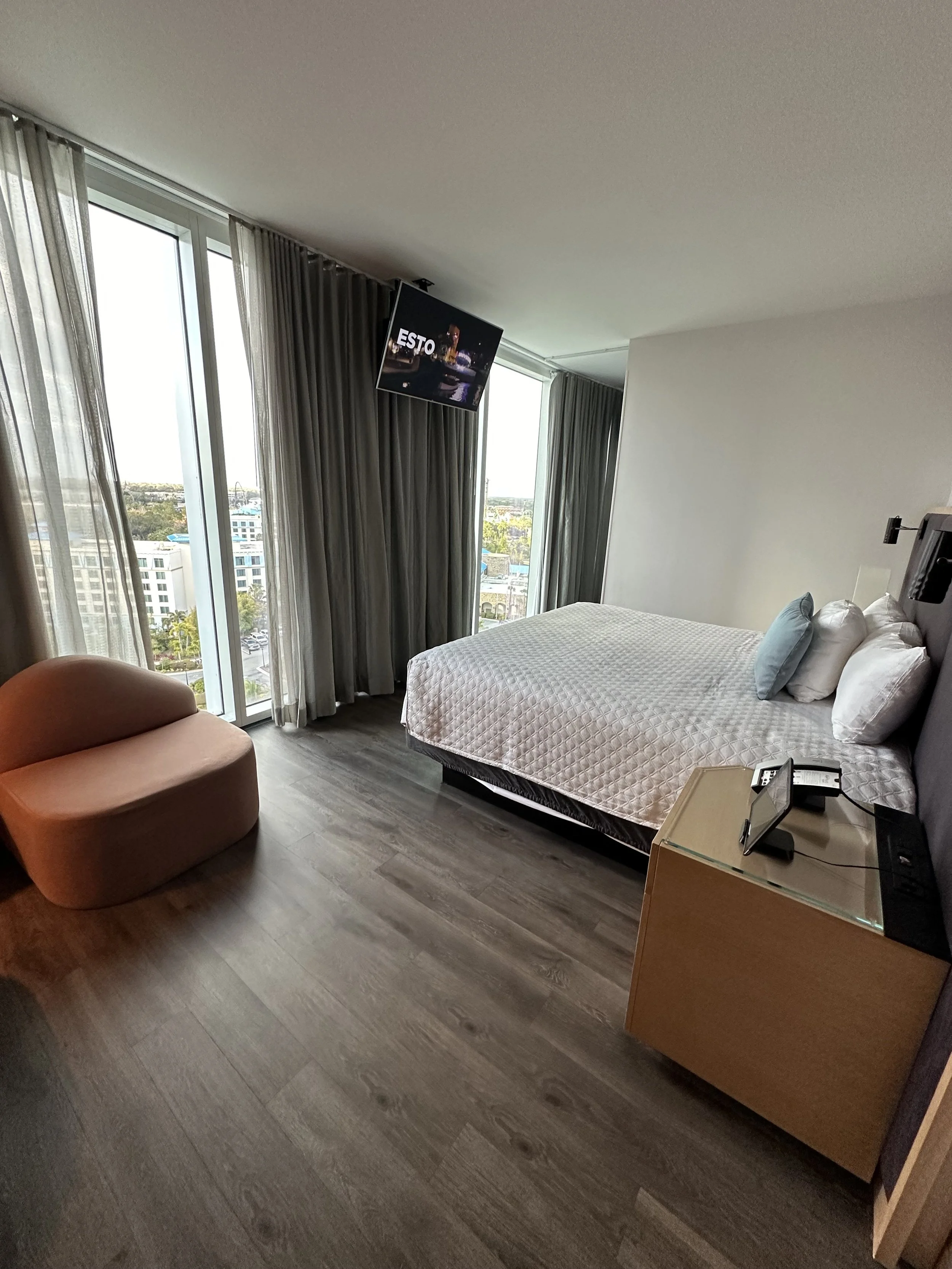A modern hotel room with a large bed, glass doors with curtains, a small orange armchair, a flat screen TV, and a nightstand with a telephone and a digital device.