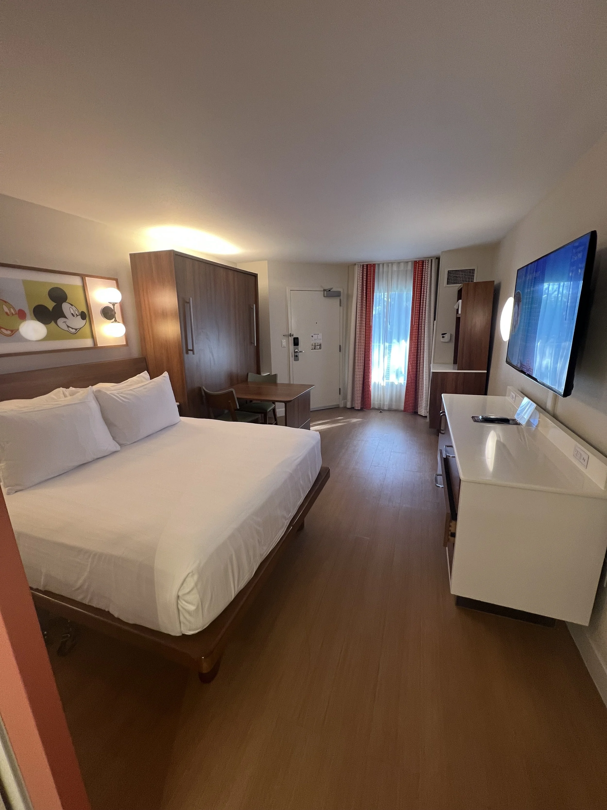 Hotel room with a bed, wardrobe, small dining table, and a large flat-screen TV mounted on the wall.