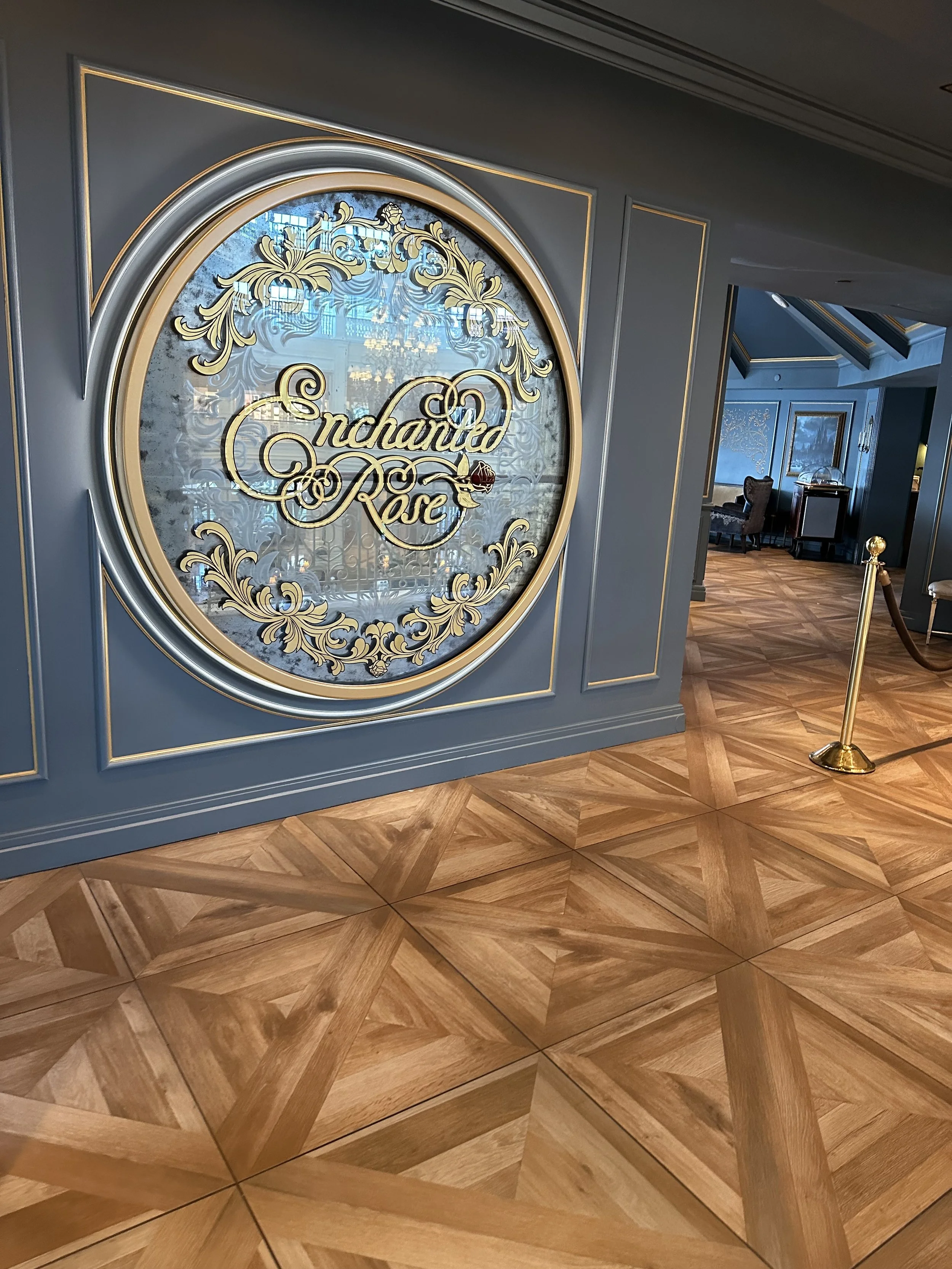 Elegant hotel lobby with a large decorative round mirror and a sign reading 'Enchanted Rose' in ornate script, framed by gold and silver accents, with patterned wooden flooring and seating area in the background.