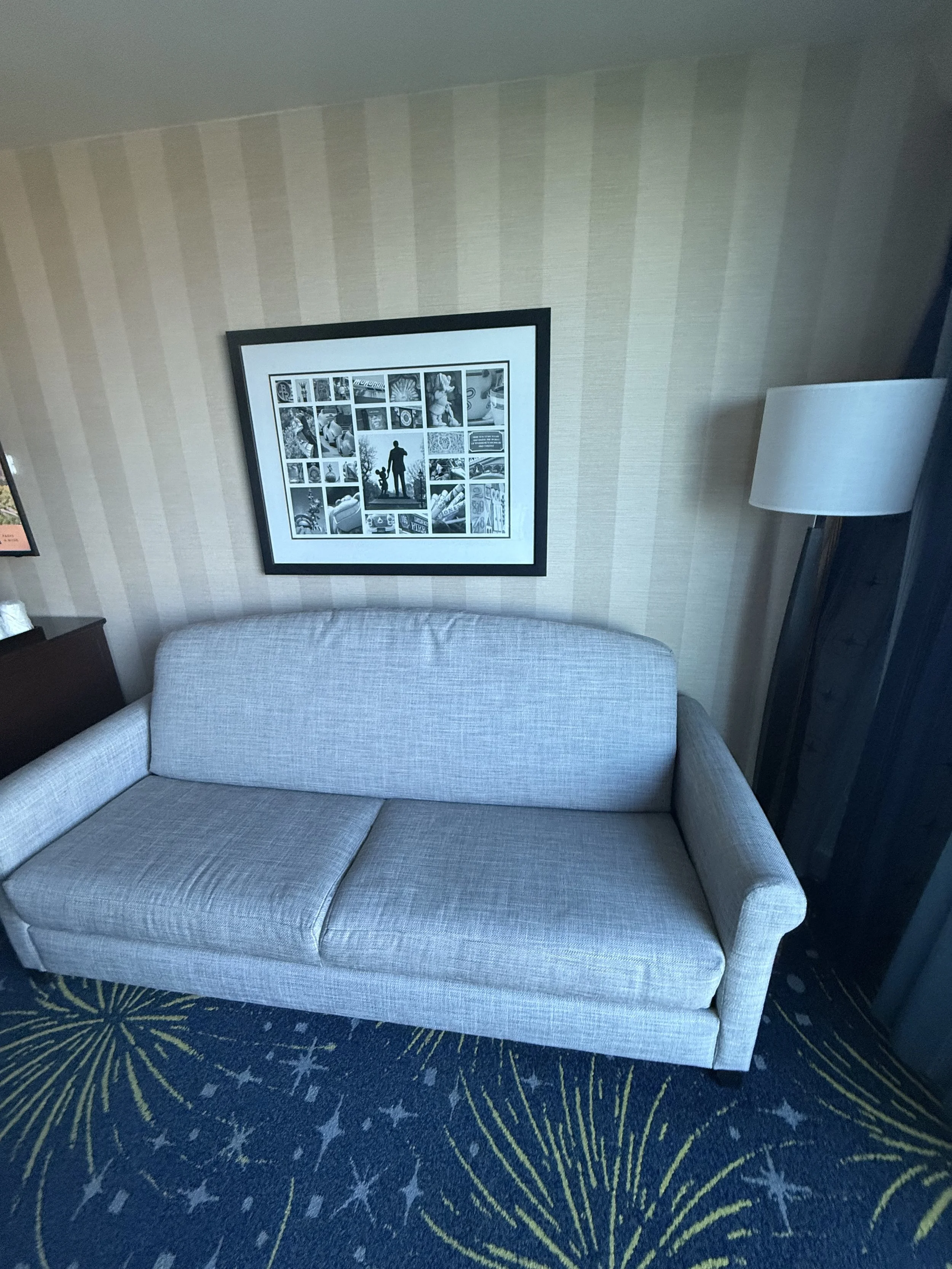 A light gray couch in a hotel room with a framed collage of black and white photographs on the wall above it. There is a wooden nightstand on the left and a tall white lamp on the right. The floor has a dark blue carpet with yellow and white starburs