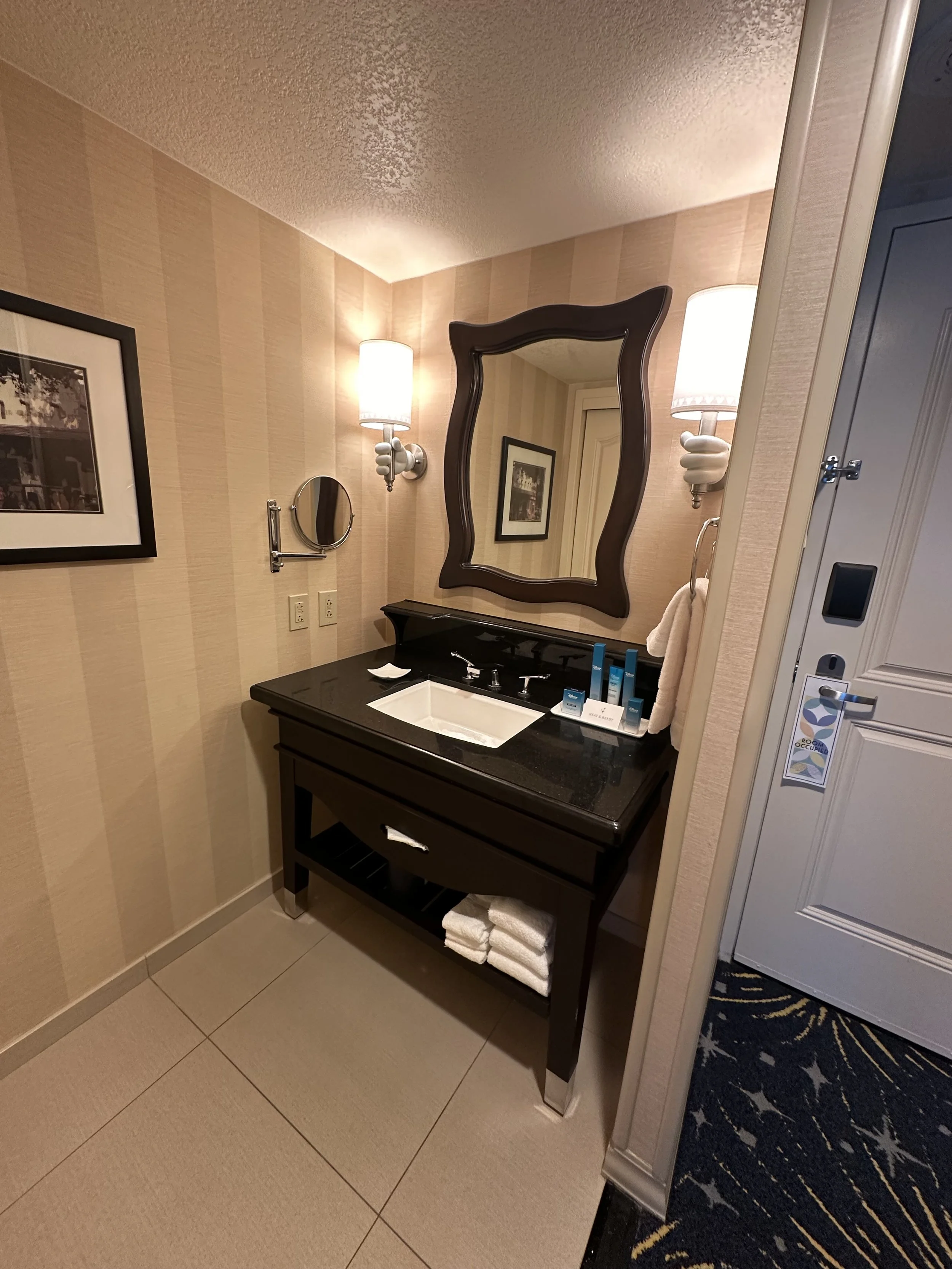 Bathroom with a black vanity, large mirror, wall-mounted light fixtures, small mirror, framed artwork, and soap and toiletries on the countertop.