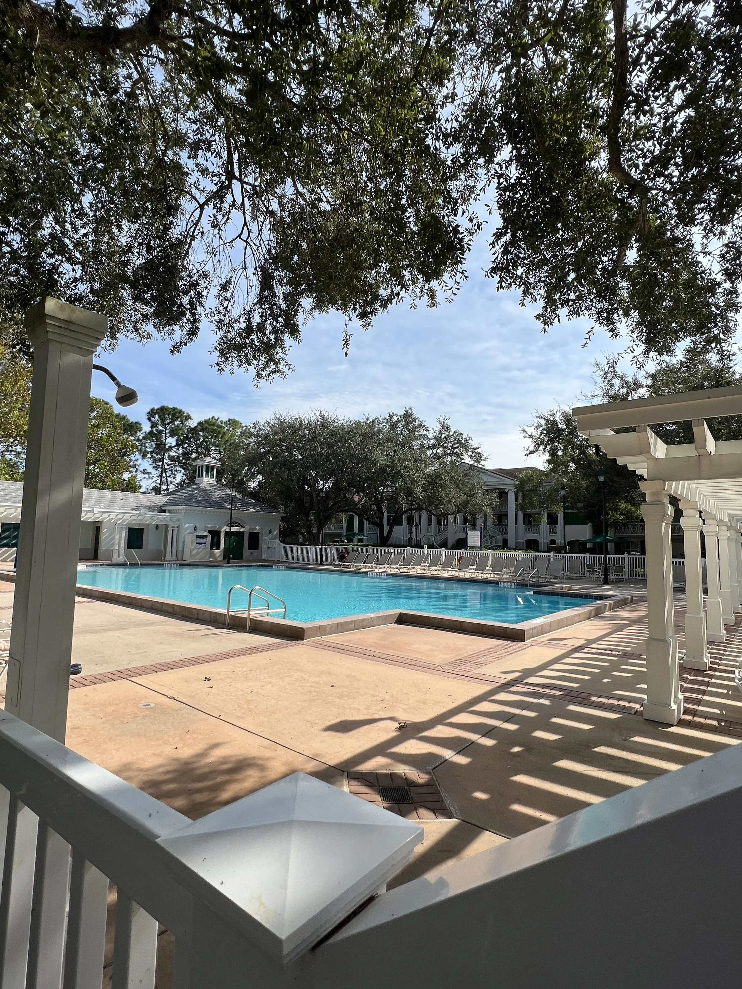Empty swimming pool surrounded by white fencing and lounge chairs, with trees and residential buildings in the background under a partly cloudy sky.