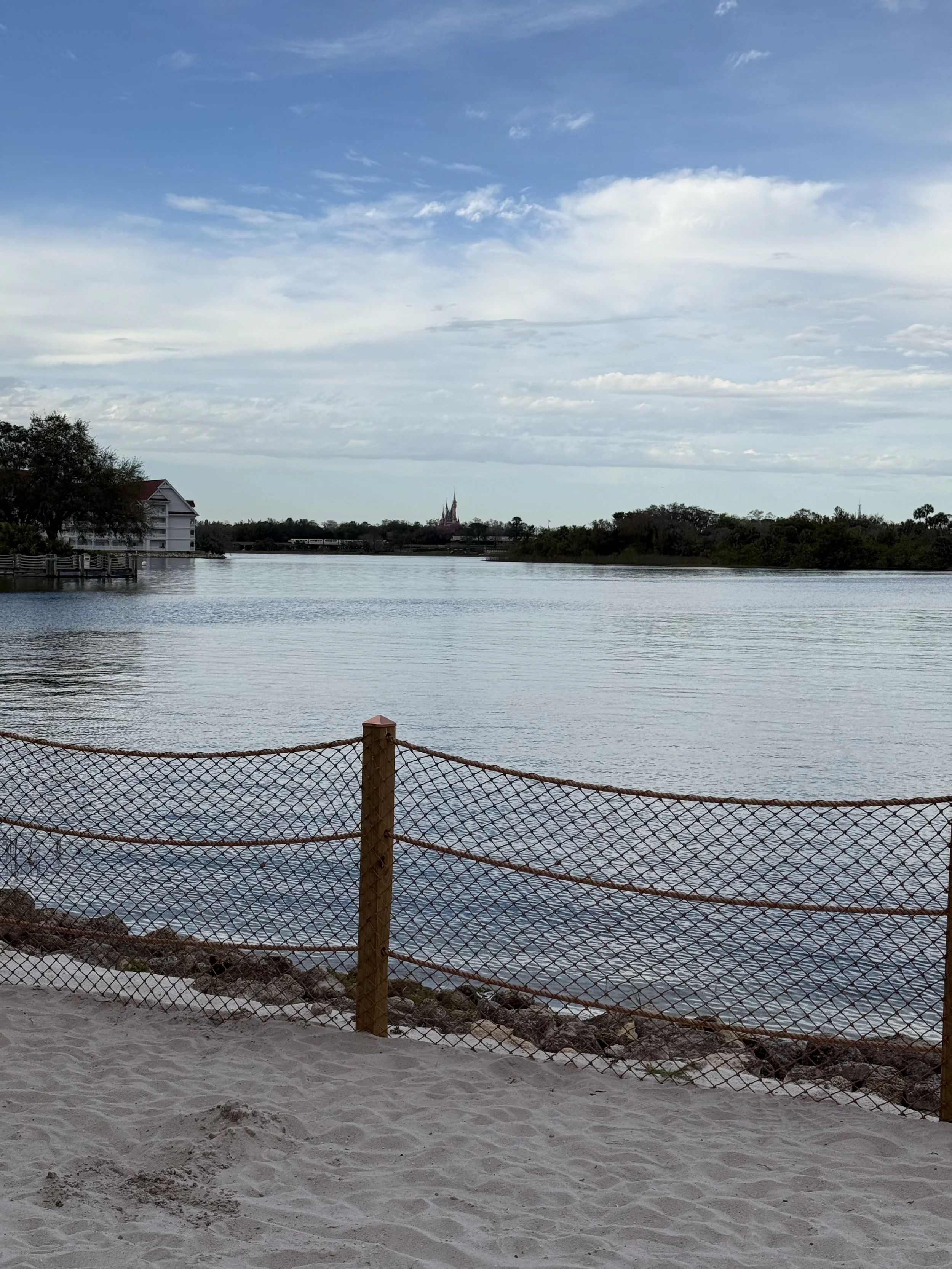 A sandy beach with a wooden and rope fence, overlooking a calm river under a partly cloudy sky, with buildings and trees along the shoreline in the distance.