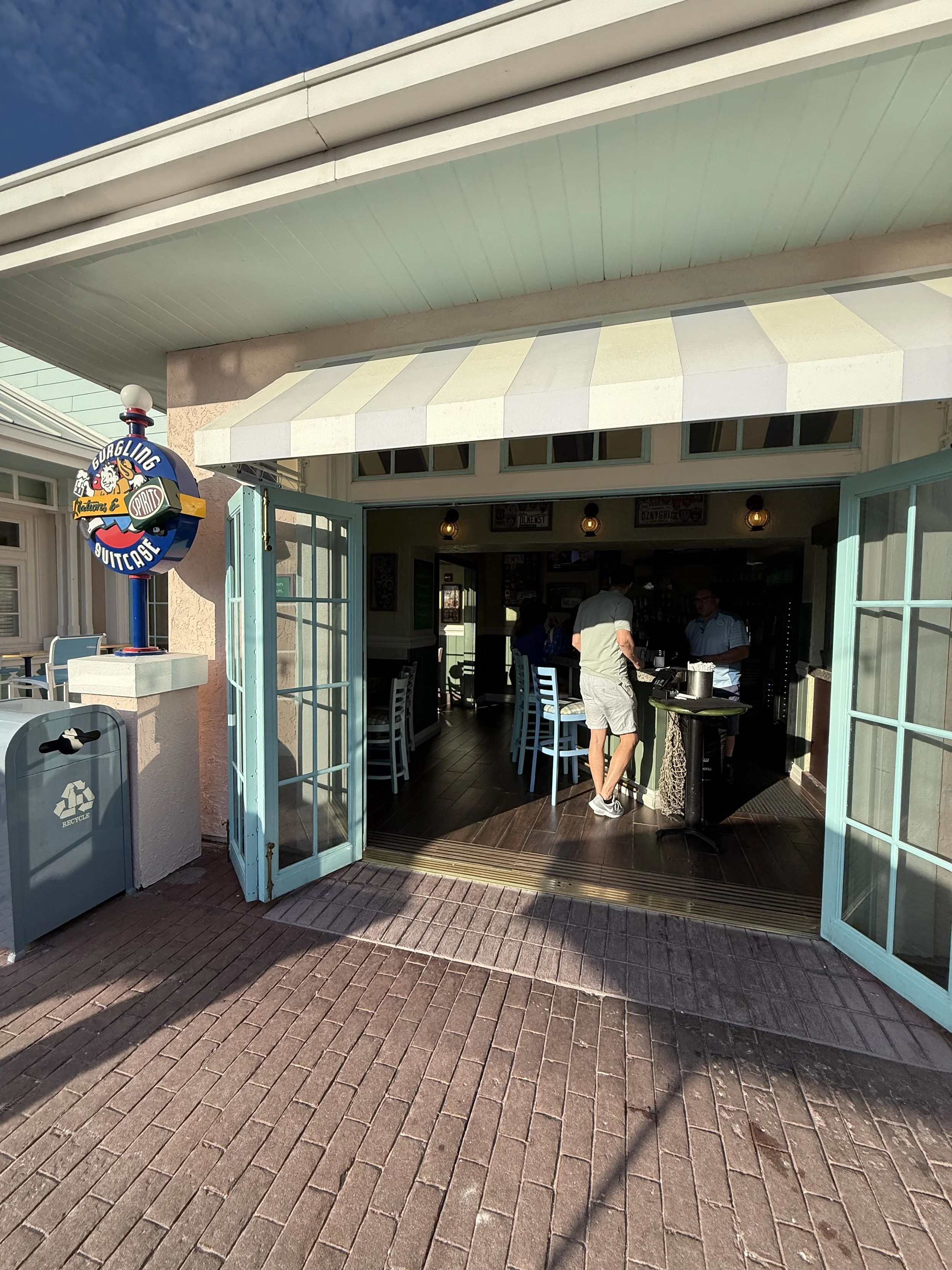 Entrance to a restaurant or cafe named Buggling Buttcafe, with people inside, open double doors, and a colorful sign outside. The sidewalk is brick, with shadows cast from nearby objects.