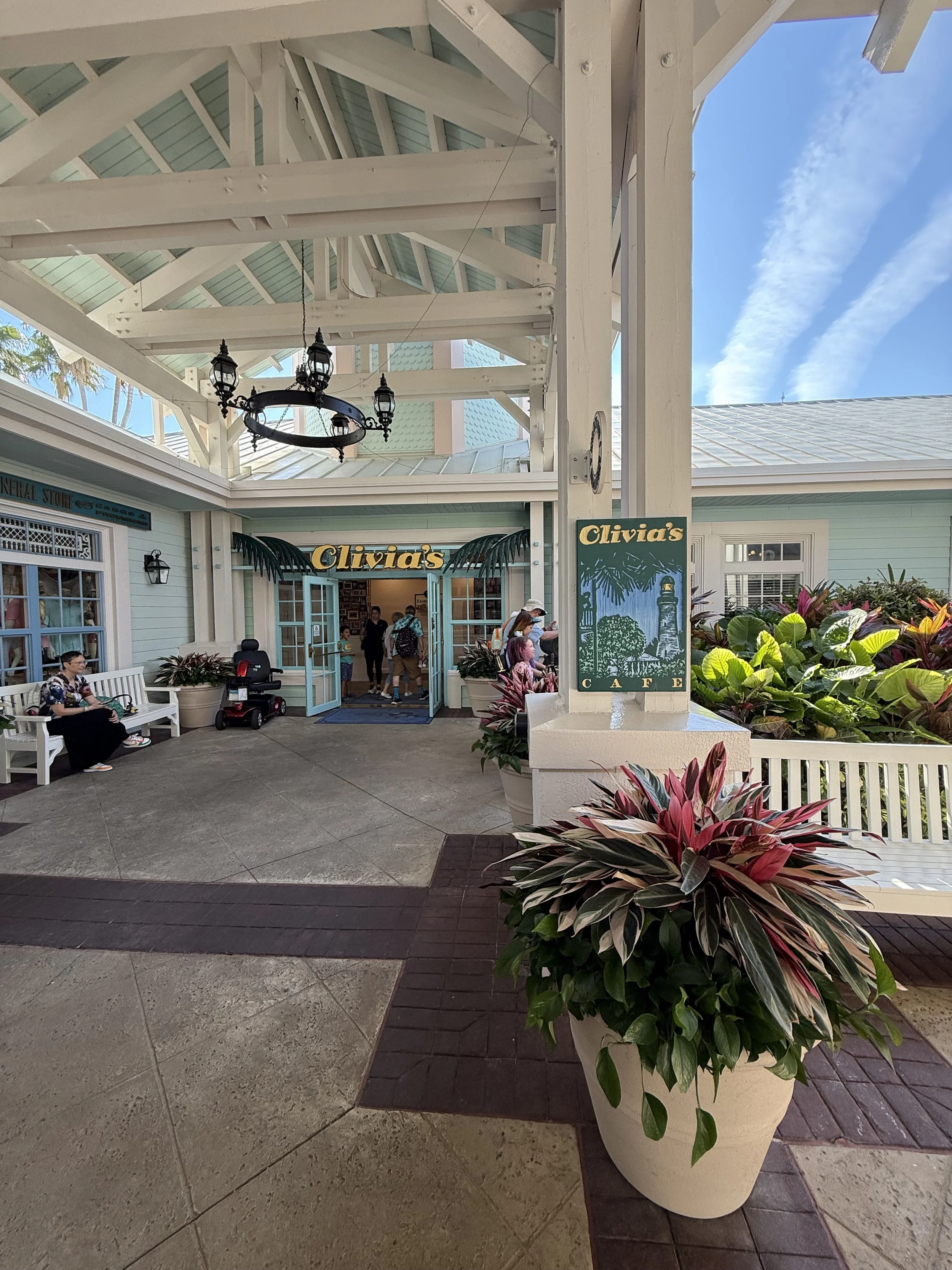 Entrance to Olivia's Cafe with potted plants, benches, and people outside under a white wooden canopy with a chandelier, blue sky, and clouds overhead.