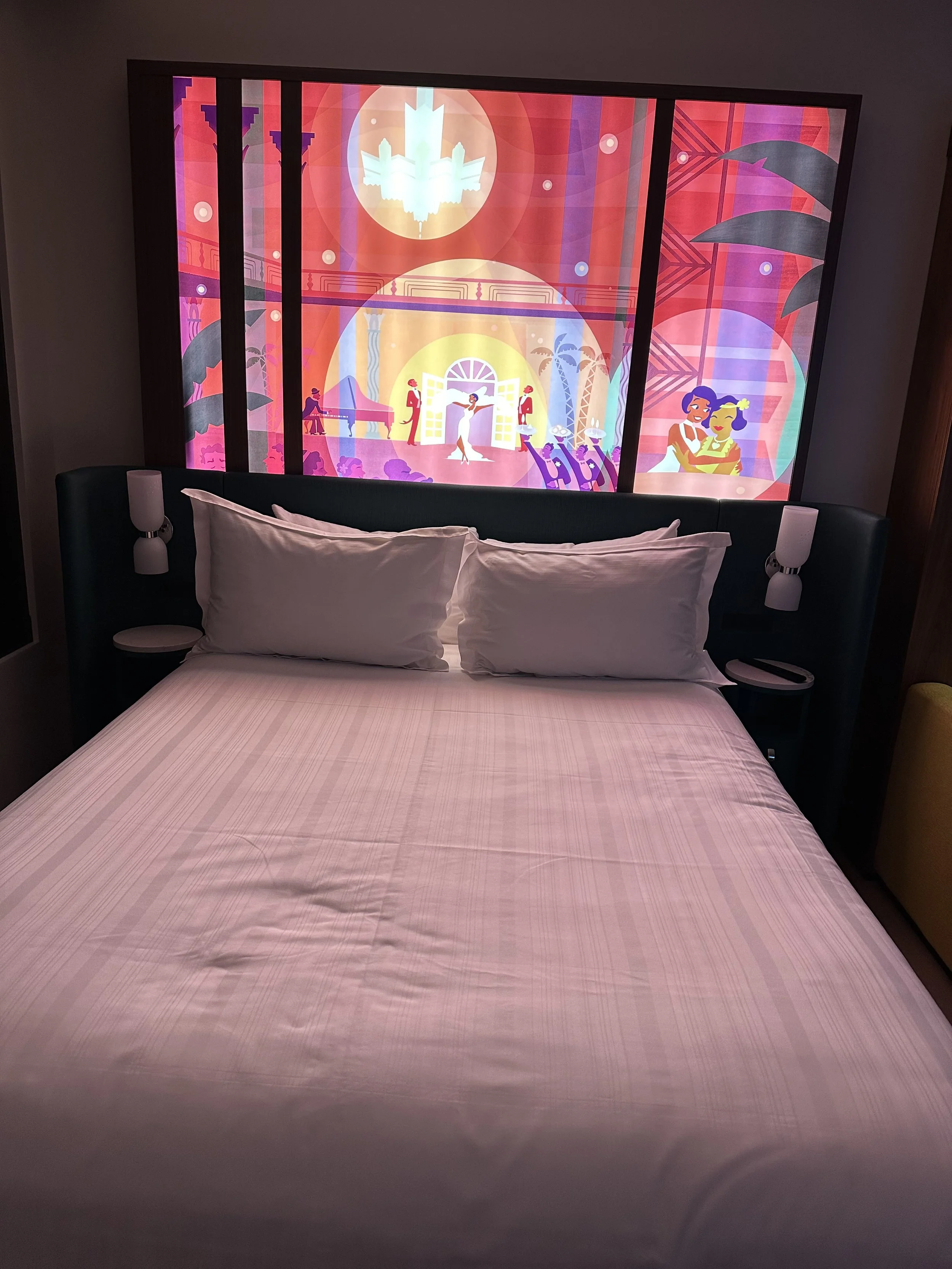 A hotel room with a neatly made bed, two pillows, and wall-mounted lamps on either side. Behind the bed, there is a colorful, animated mural depicting a lively scene with people dancing, playing music, and socializing, set in a vibrant, stylized indo