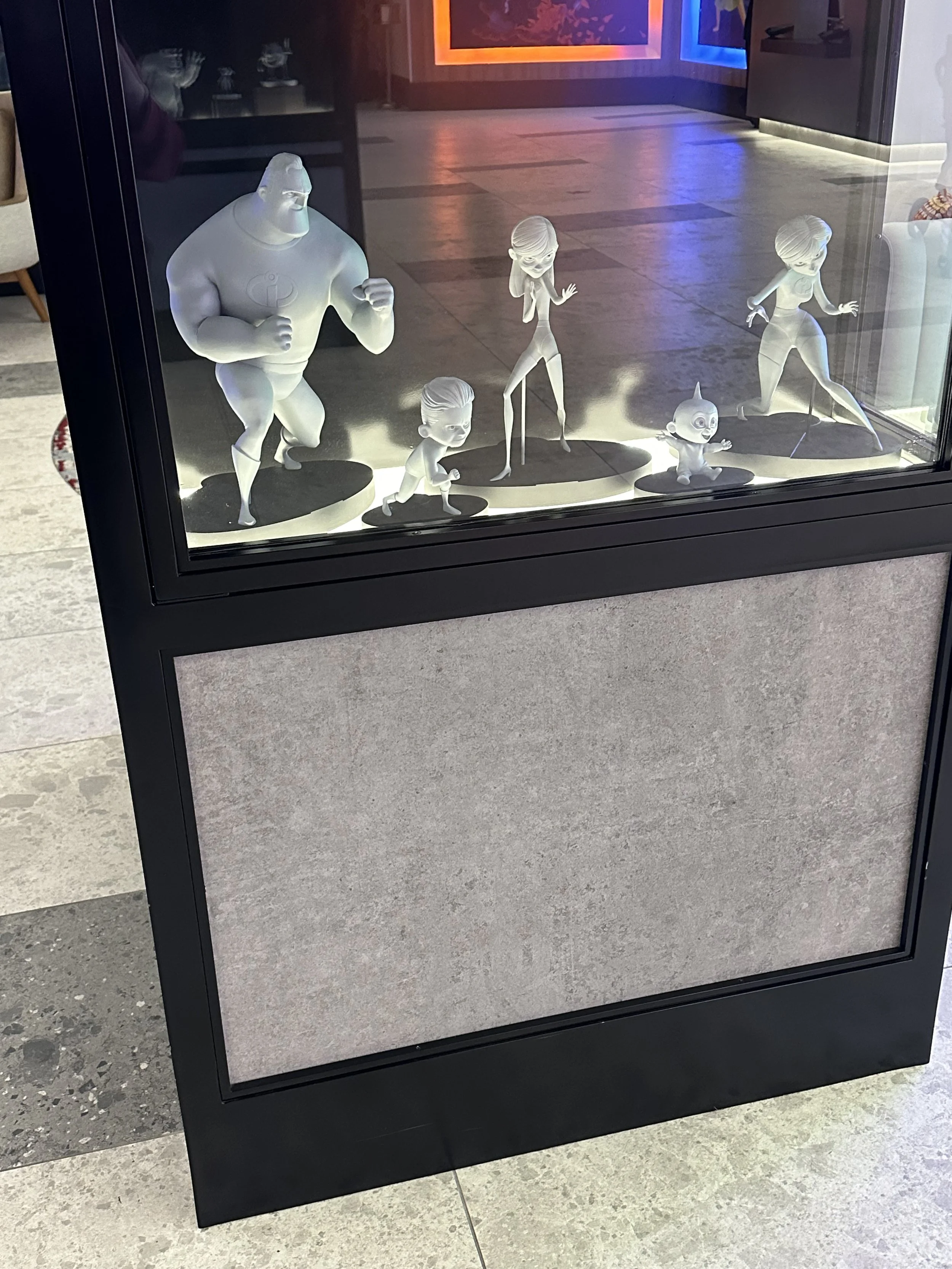 Display case with five cartoon-like character figurines, including a large muscular man, a small girl, a tall woman, a baby, and a woman in a bodysuit, in a brightly lit room with colorful wall lights.