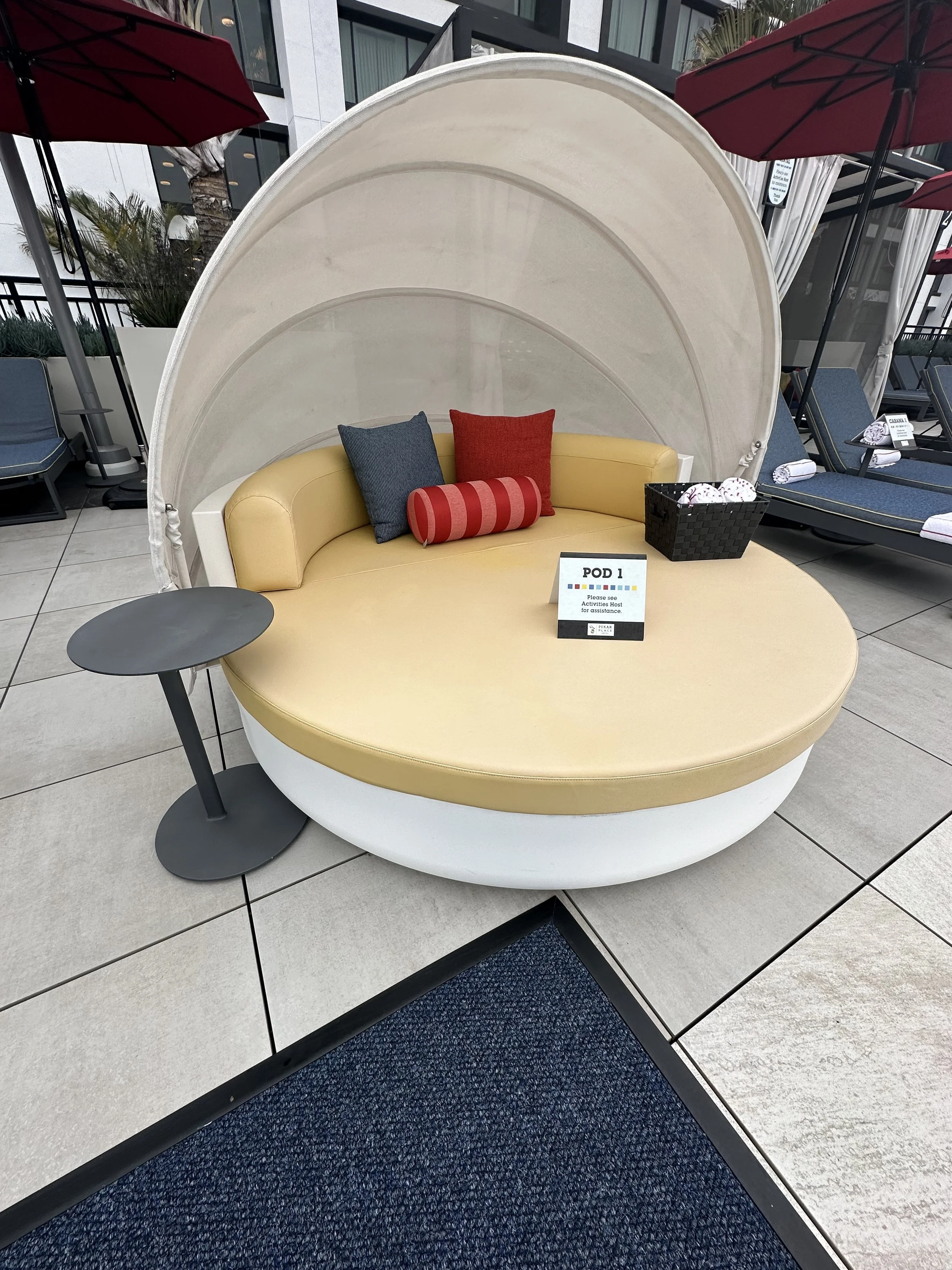 A circular sunbed with a beige cushion and a semi-circular white canopy, accented by three pillows in blue, red, and red with stripes, situated on a tiled outdoor patio with lounge chairs and umbrellas.