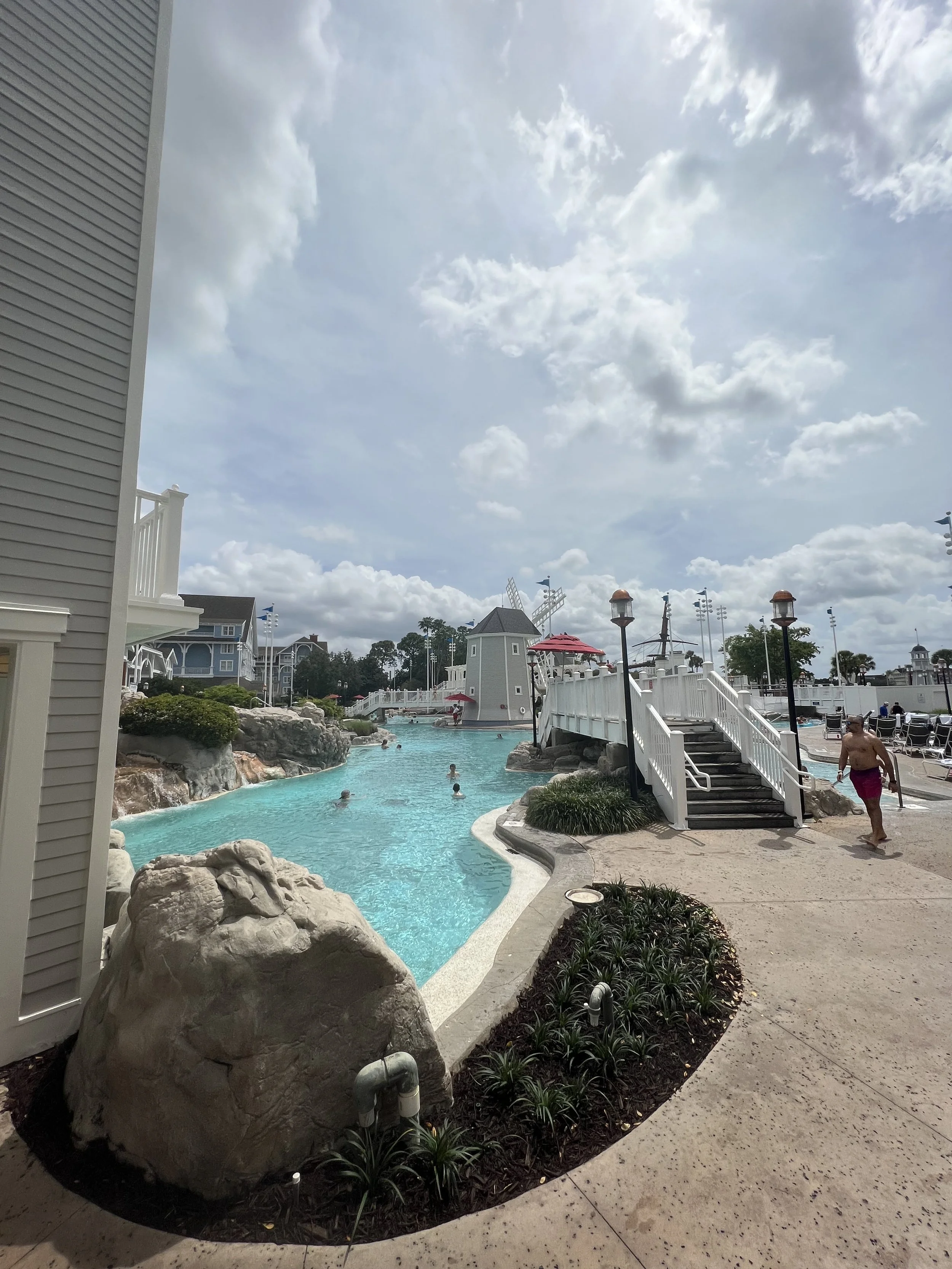 A pool area with clear blue water, rocks, and surrounding plants, with a white staircase leading to a small white building with a windmill decoration, several people swimming in the pool, and others walking nearby under a partly cloudy sky.