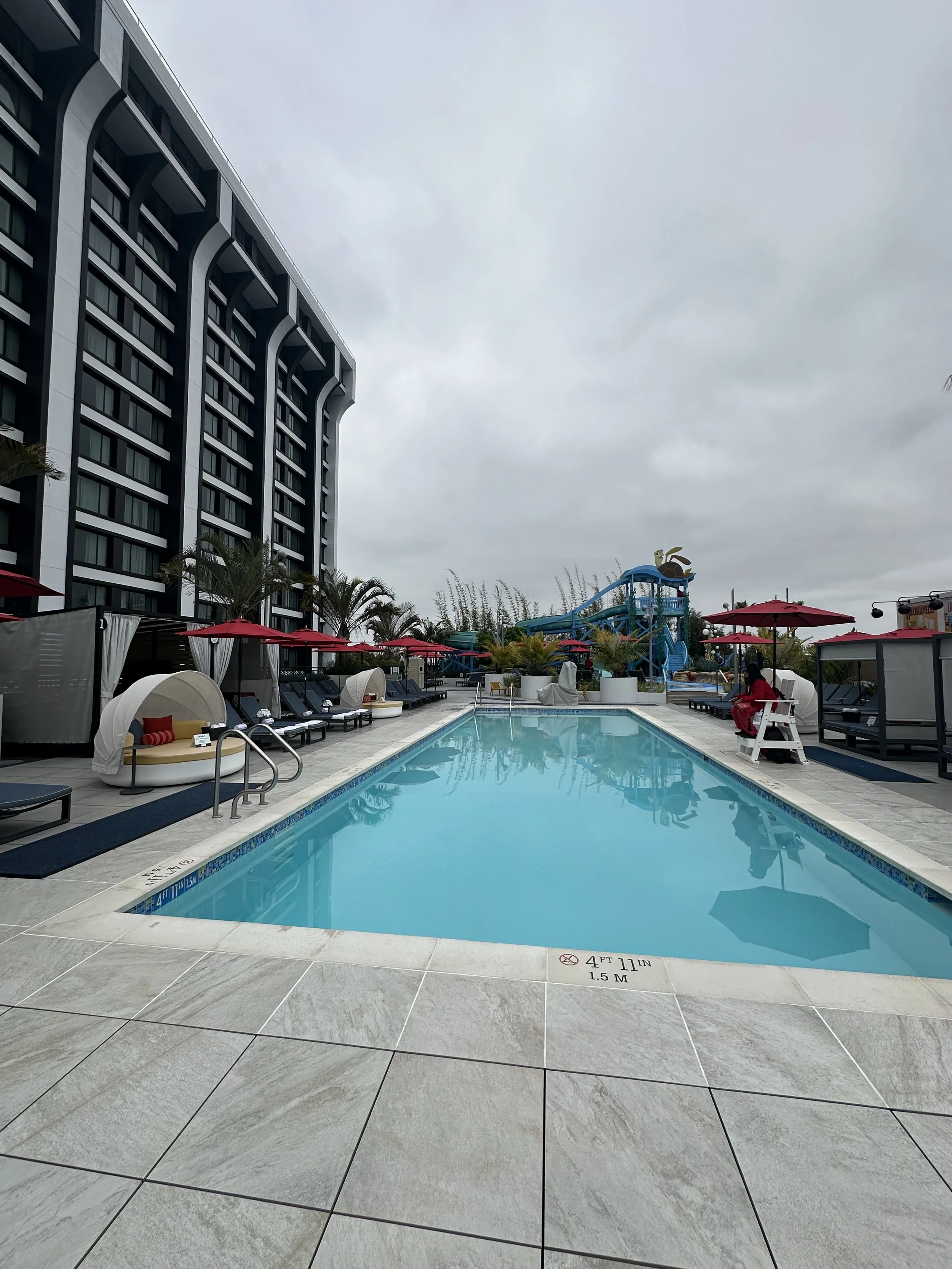 Empty outdoor hotel pool with lounge chairs and red umbrellas, water slide in the background, and a tall building on the left, under a cloudy sky.