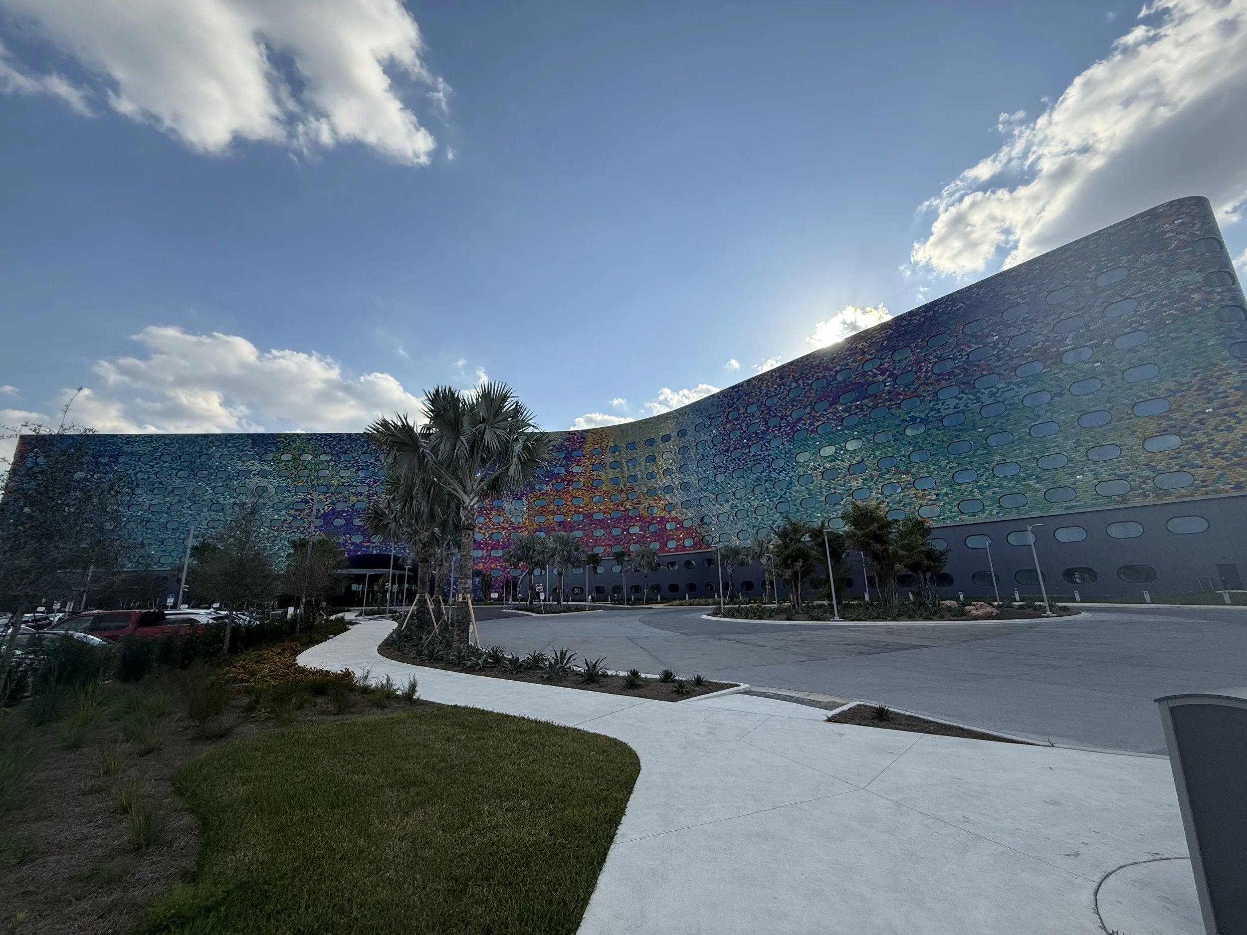 A large, colorful, curved building with a mosaic-like facade under a partly cloudy sky, surrounded by palm trees and a parking lot.