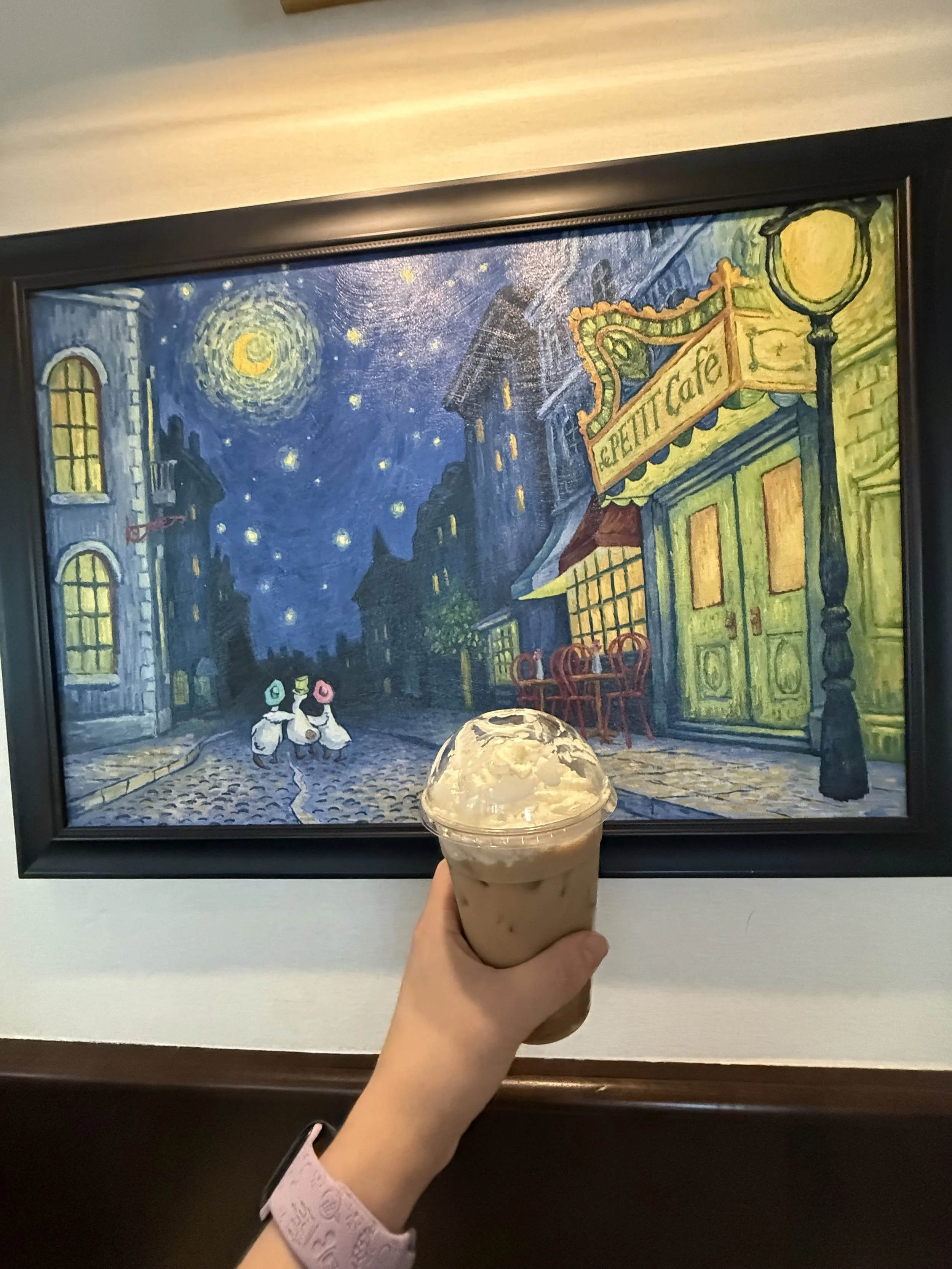 A hand holding a cold coffee drink in front of a framed painting of a nighttime street scene with the moon, stars, and buildings, including a cafe.